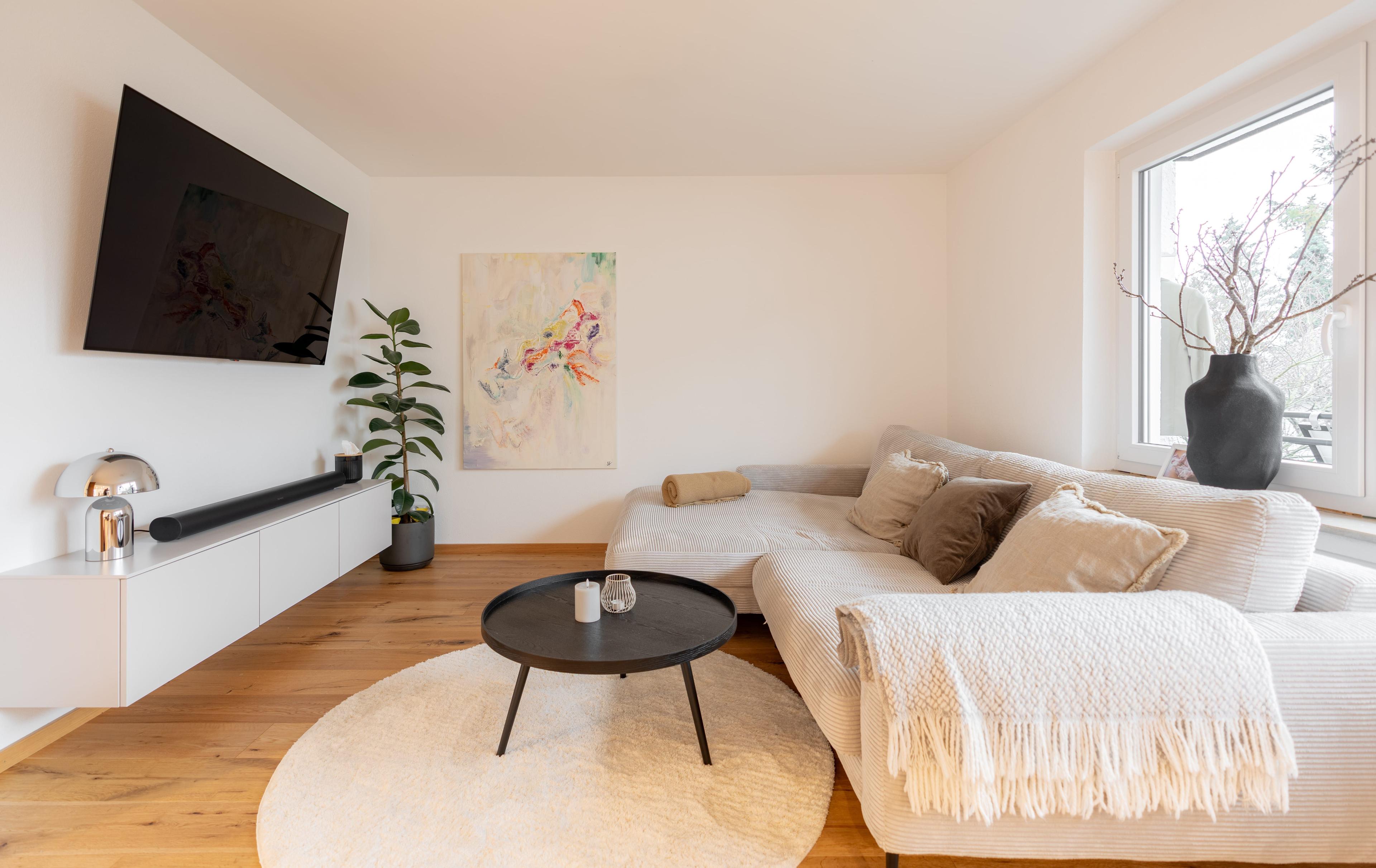 Gemütlicher Wohnbereich mit beigem Ecksofa, schwarzem Couchtisch, weißem TV-Board, Zimmerpflanze und abstraktem Wandbild.
