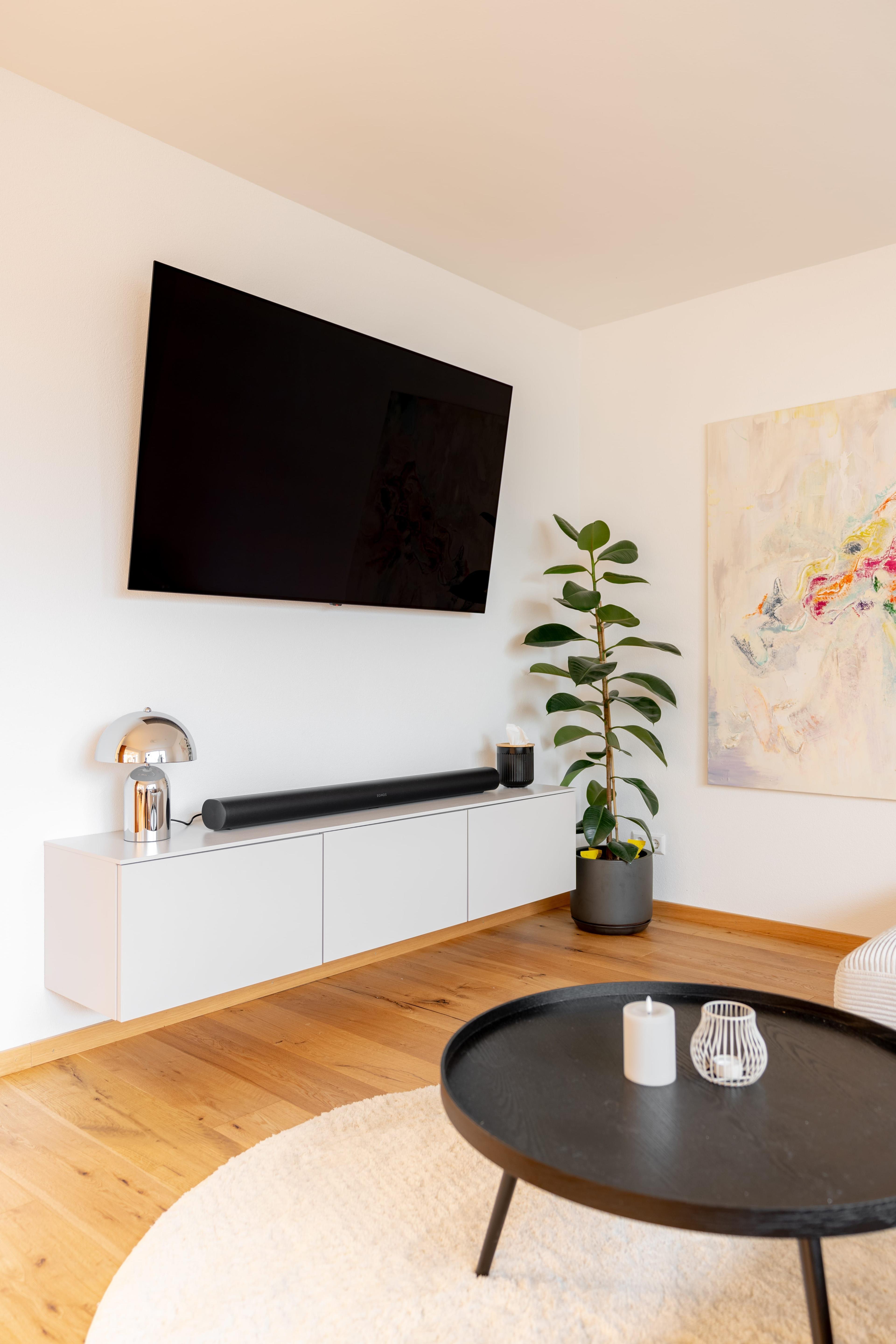 TV-Wand mit weißem hängendem Sideboard, schwarzer Soundbar, Chrom-Tischlampe, Zimmerpflanze und Wandfernseher.