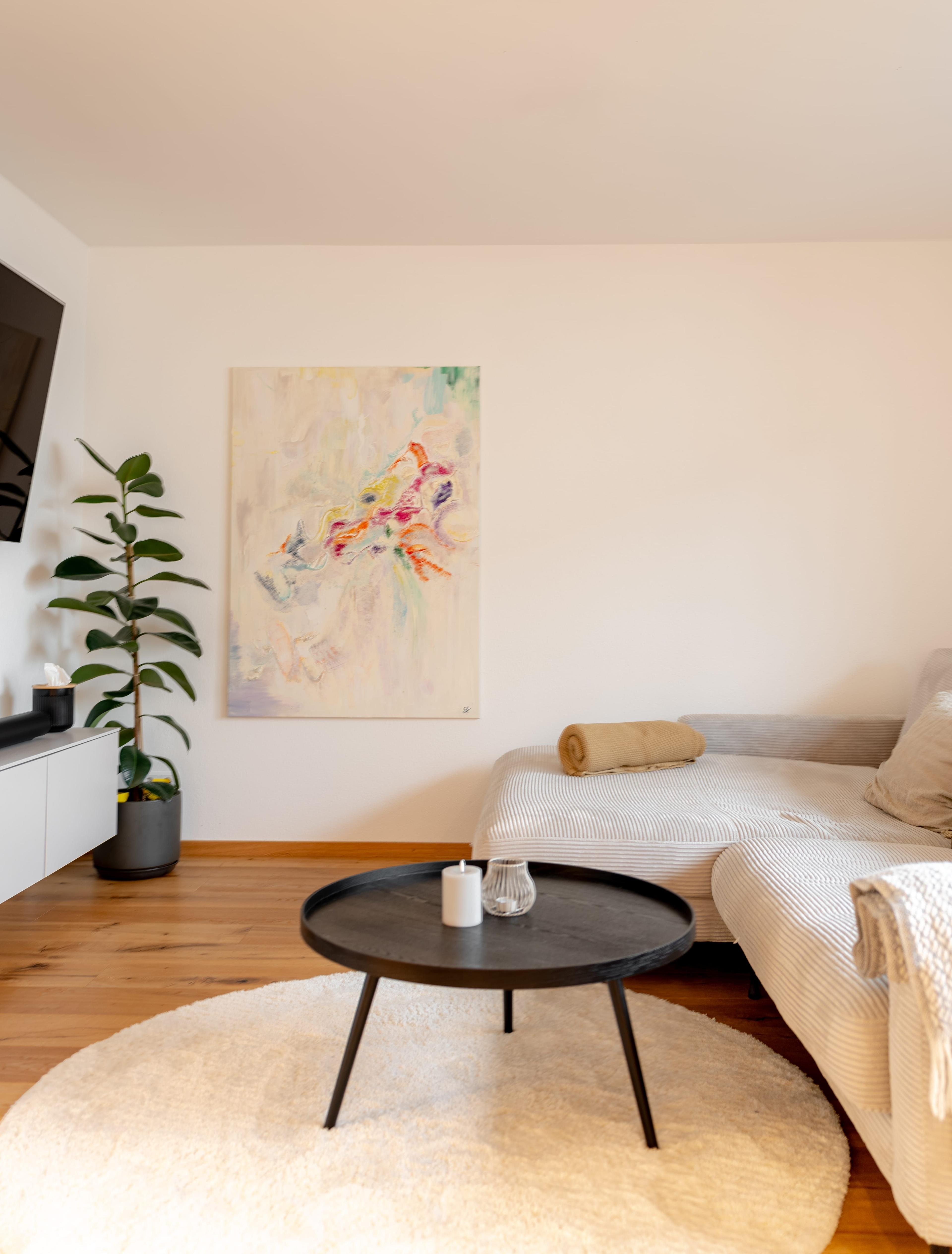 Sitzecke mit beigem Sofa, abstraktem Kunstwerk, schwarzem Couchtisch, Zimmerpflanze und hellem rundem Teppich.