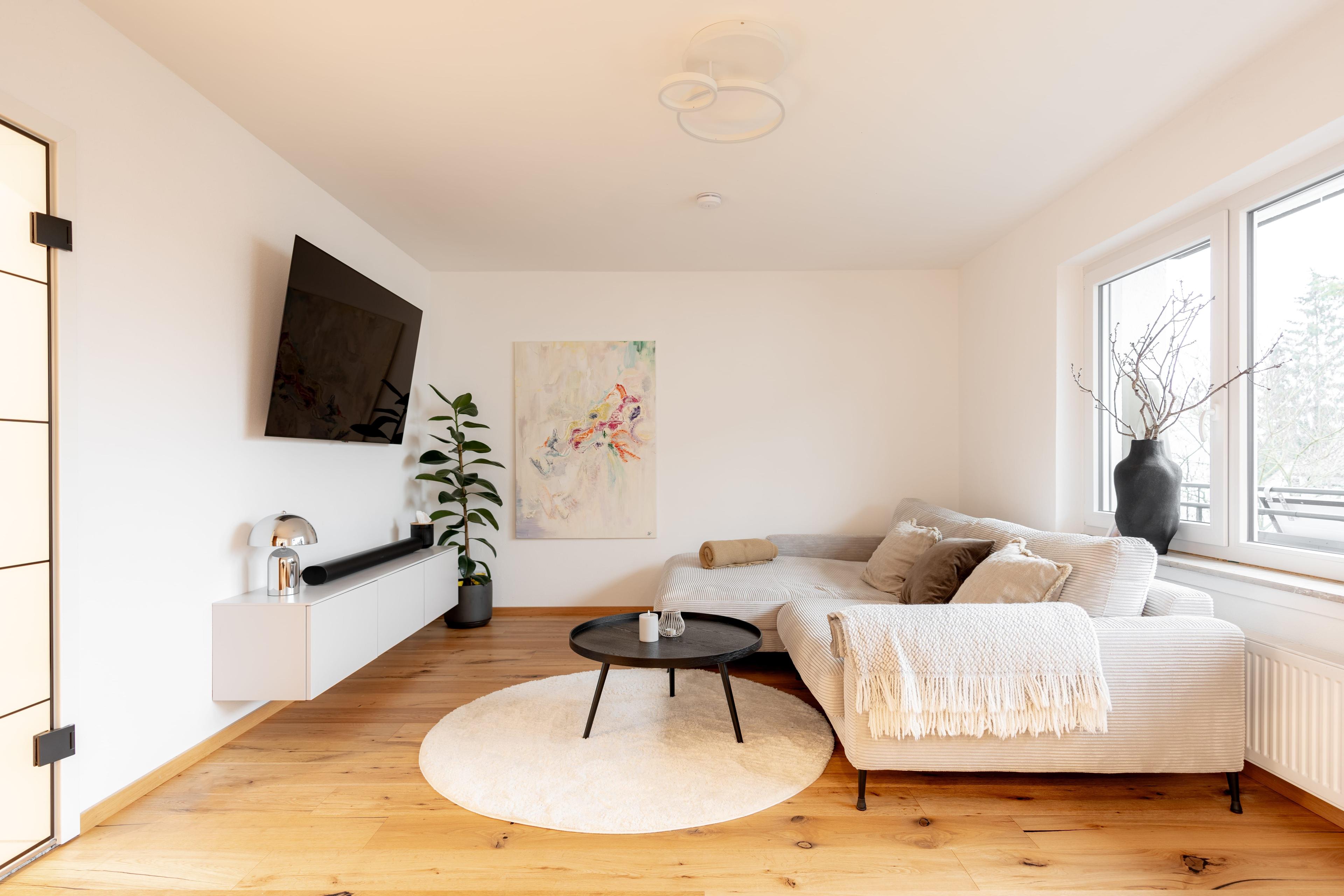 Wohnzimmer-Gesamtansicht mit Cord-Sofa, TV-Board, Designer-Pendelleuchte, großen Fenstern und warmem Holzparkett.