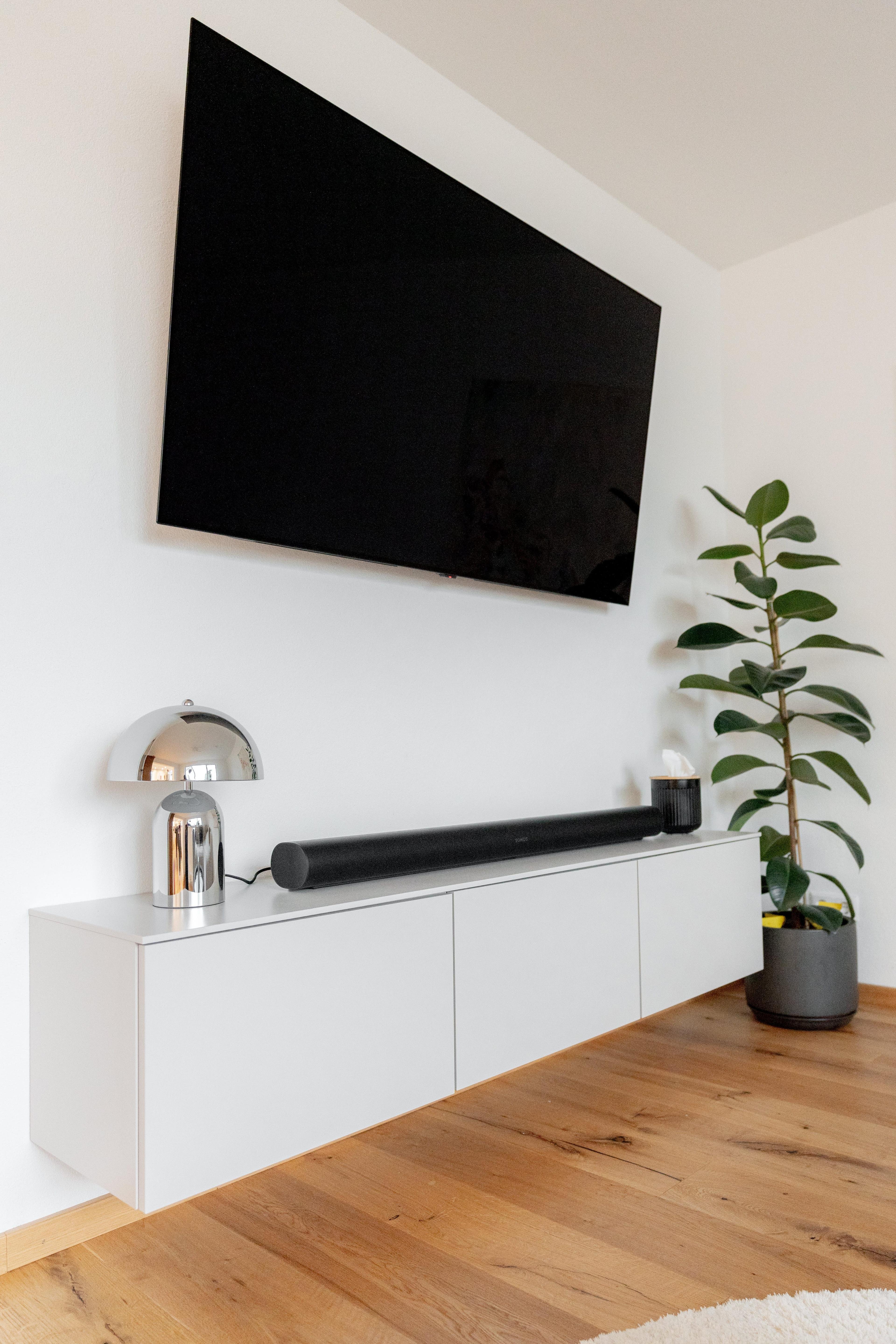 Detailansicht TV-Bereich mit wandhängendem weißen Sideboard, schwarzer Soundbar, Designerlampe und Gummipflanze.
