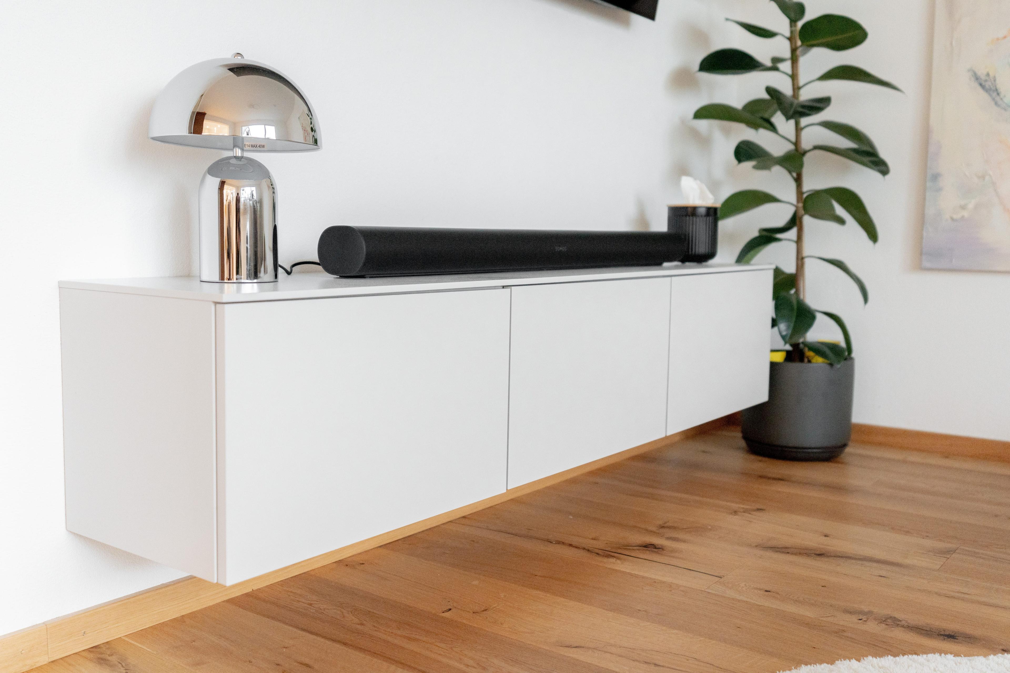 Nahaufnahme TV-Lowboard mit Chrom-Pilzlampe, schwarzer Soundbar, weißen Fronten und Zimmerpflanze auf Eichenparkett.