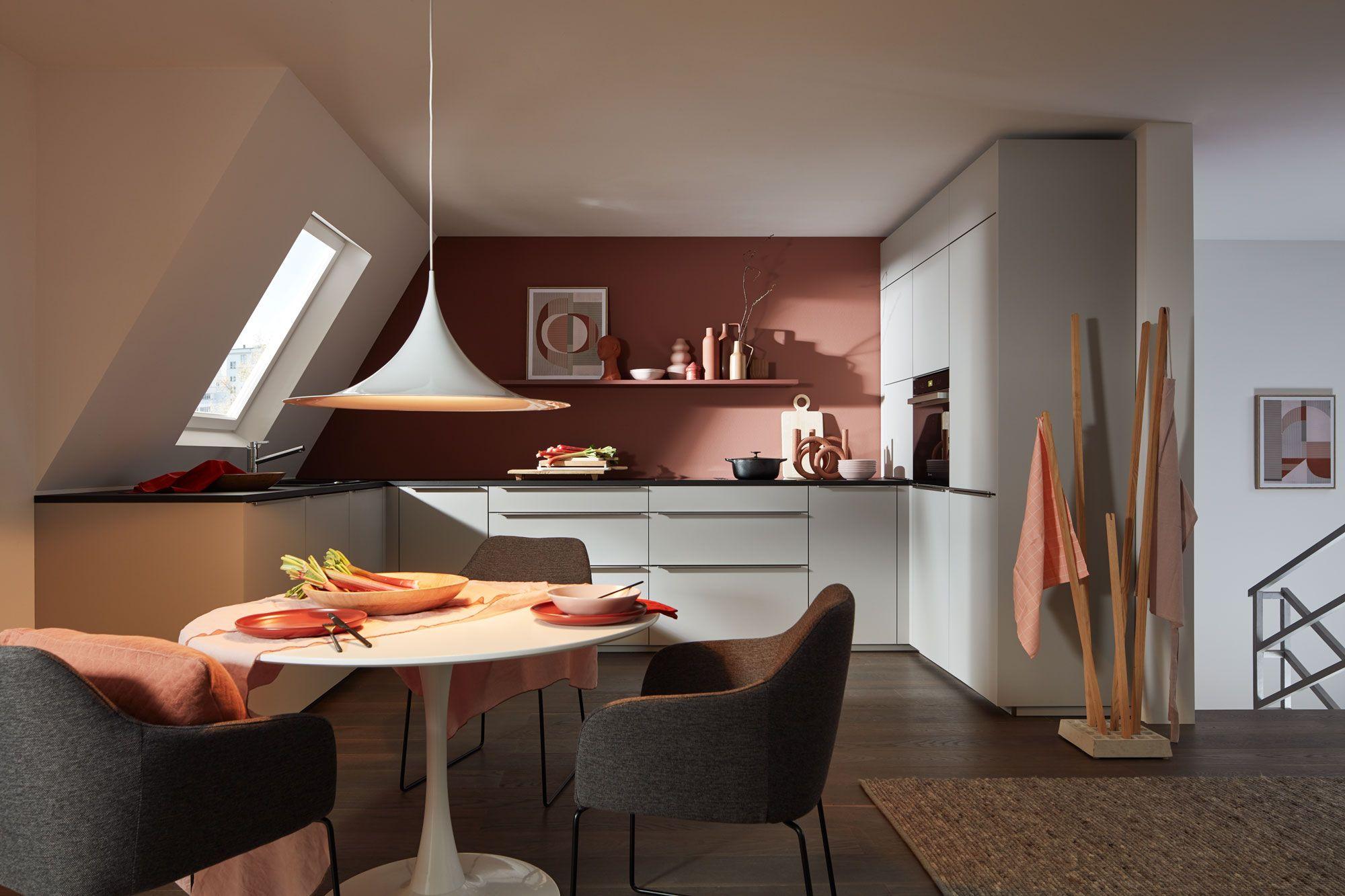 Moderne Küche mit rosa Wandfarbe, weißem Küchenblock und Esstisch mit eleganten Stühlen, ergänzt durch dekorative Objekte und eine stilvolle Pendelleuchte.
