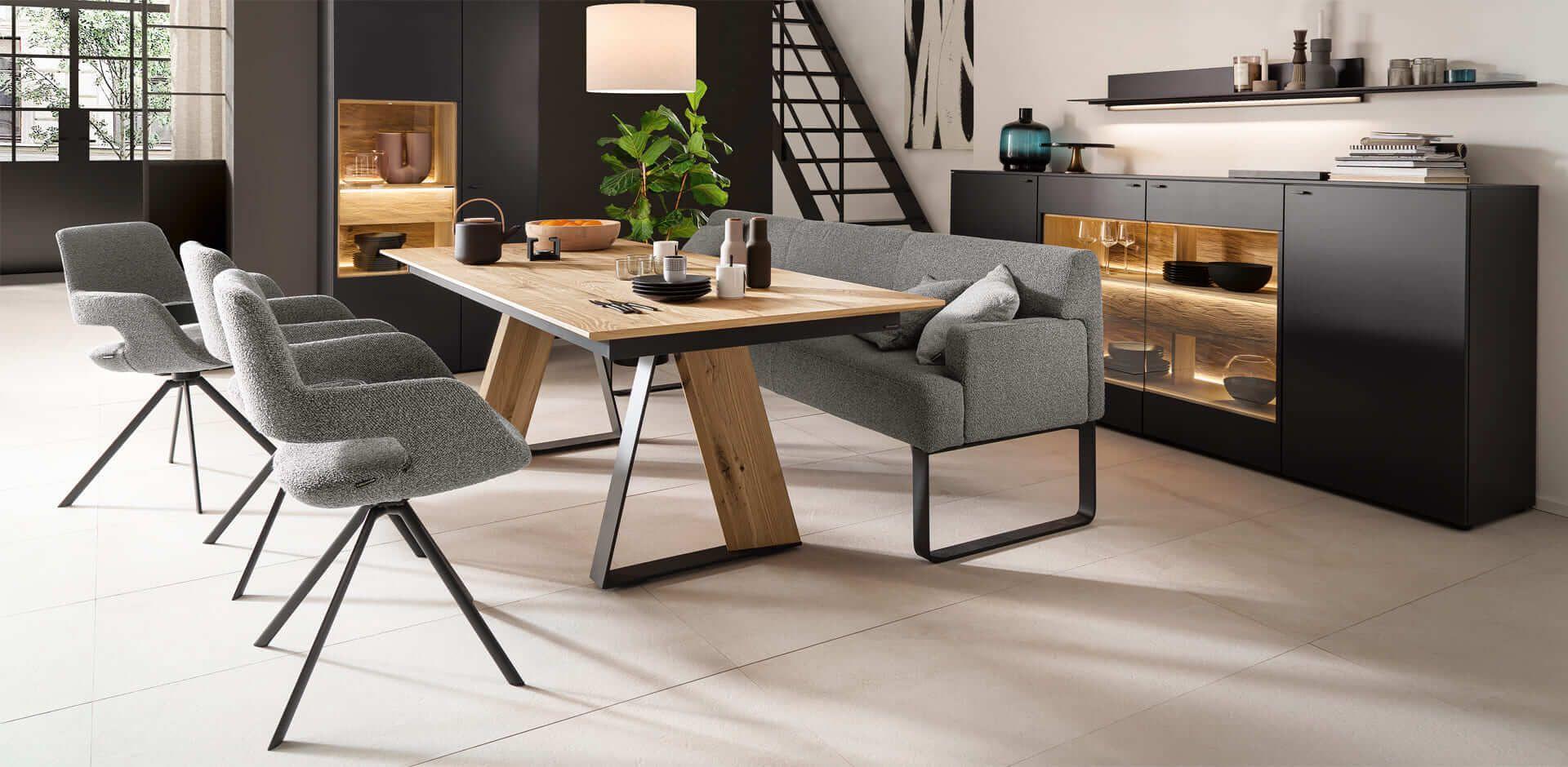 Moderner Esstisch mit Holzplatte und Metallgestell, graue Polsterstühle und Eckbank vor anthrazitfarbenem Sideboard mit LED-Beleuchtung in offenem Wohnbereich.
