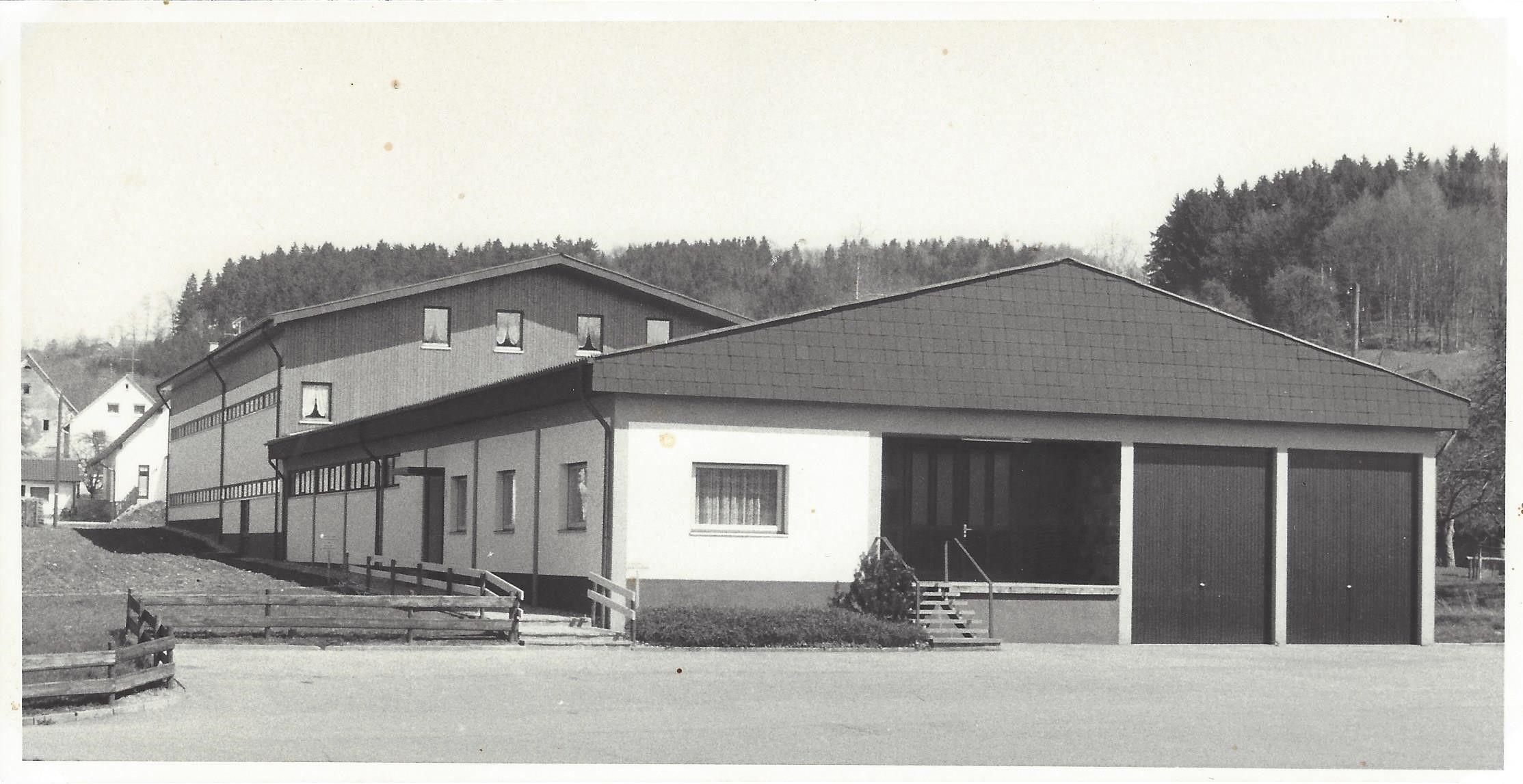 Historische Schwarz-Weiß-Fotografie eines modernen Industriegebäudes mit Flachdach und großen Toren vor bewaldeter Bergkulisse, vermutlich 1960er Jahre.