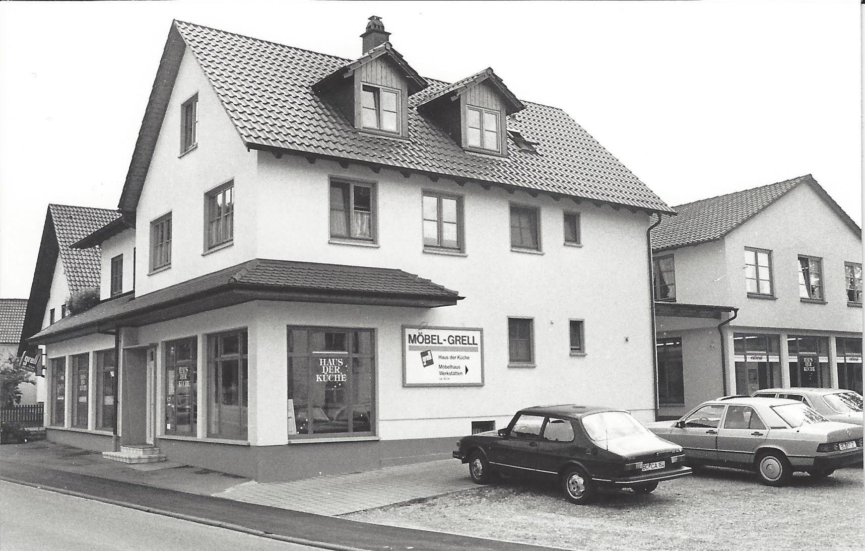 Historische Schwarz-Weiß-Fotografie eines Möbelgeschäfts "Möbel-Grell" mit "Haus der Küche" in einem zweistöckigen Gebäude mit Dachgauben und geparkten Autos der 1970er/80er Jahre davor.