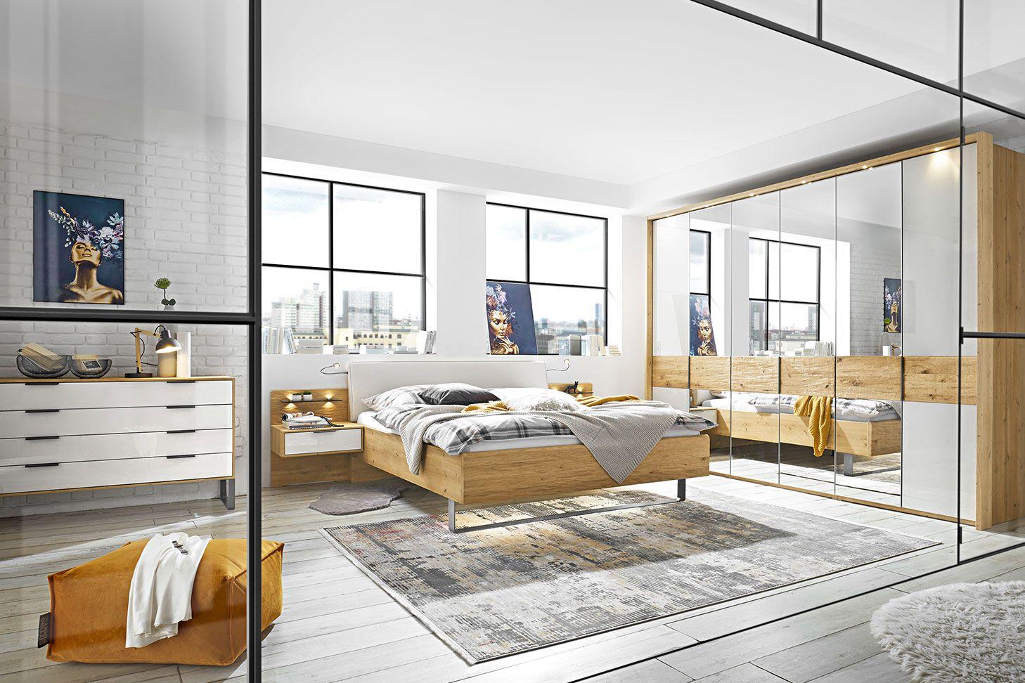 Loft-Schlafzimmer mit Holzbett, großem Spiegelschrank, weißer Backsteinwand, Industriefenstern und bunten Kunstwerken an der Wand.