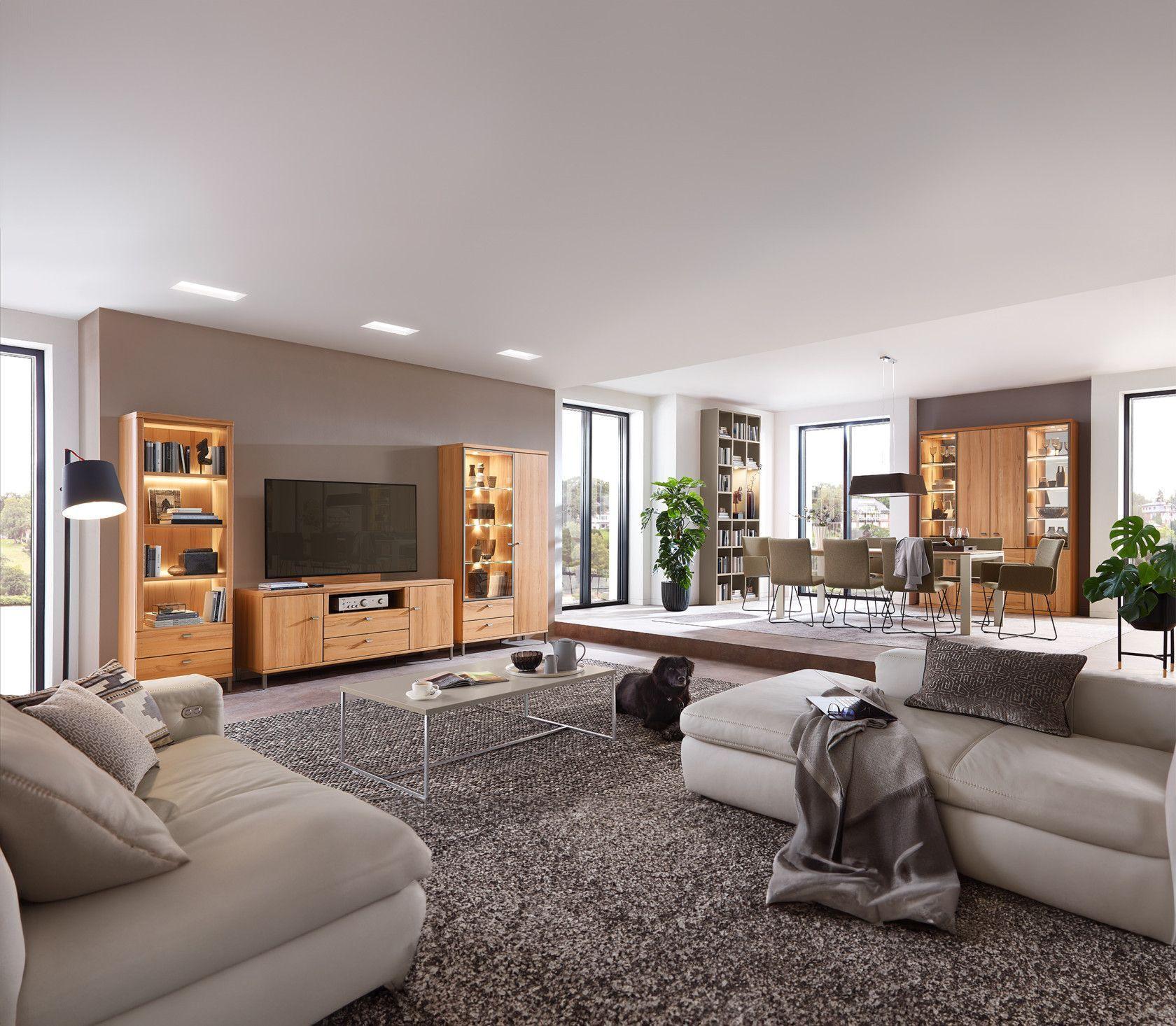 Offener Wohnbereich mit beigen Ledersofas, Holz-TV-Wand mit beleuchteten Vitrinen, schwarzem Hund auf grauem Teppich und Essbereich mit Bücherregal im Hintergrund.