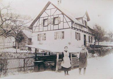 Historische Schwarz-Weiß-Fotografie zeigt ein Fachwerkhaus im Winter mit zwei Personen in traditioneller Kleidung vor einem Holzzaun im Schnee.