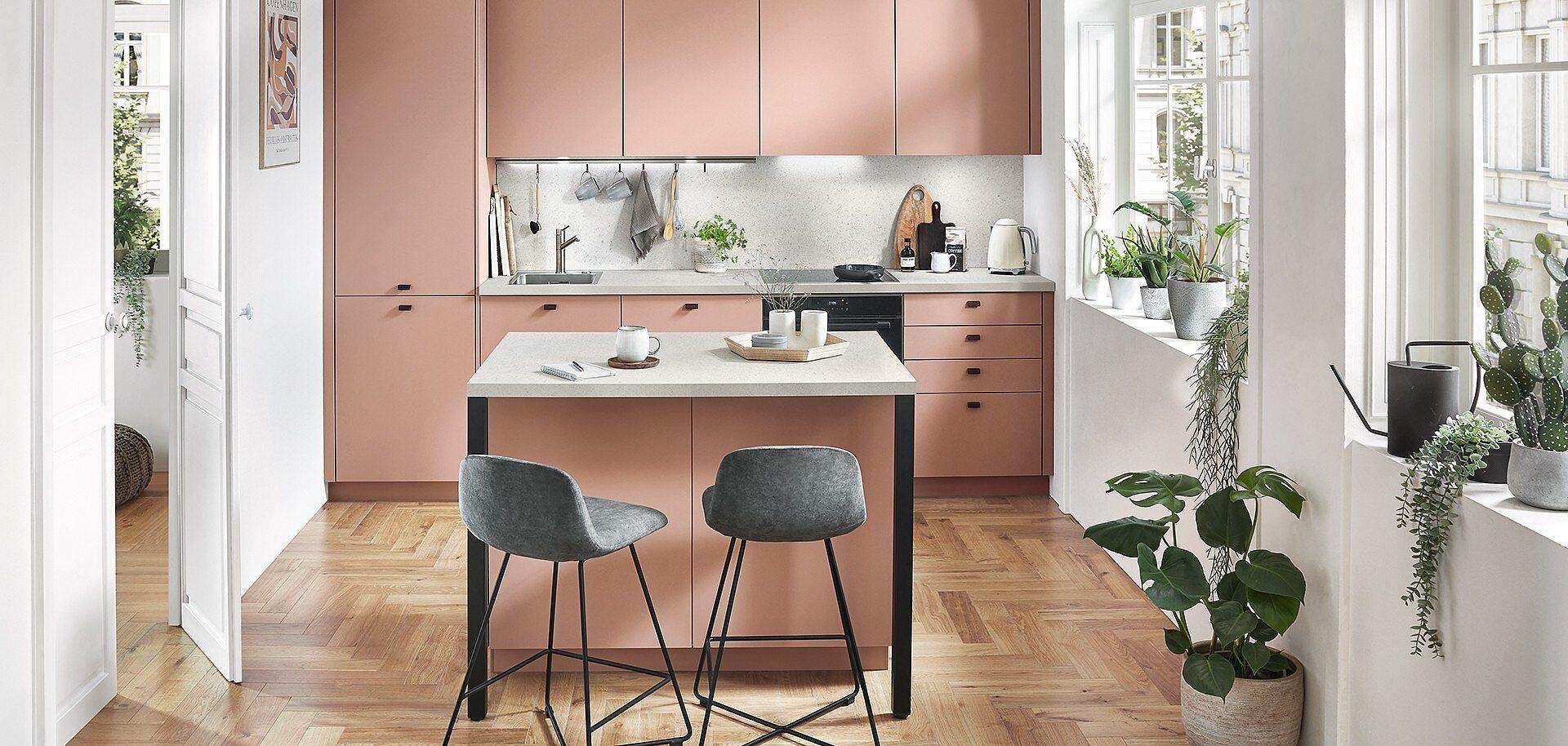 Moderne Kücheninsel in Rosa-Kupfer-Optik mit weißer Arbeitsplatte, grauen Barhockern und grünen Pflanzen in einem hellen Raum mit Holzboden und großen Fenstern.