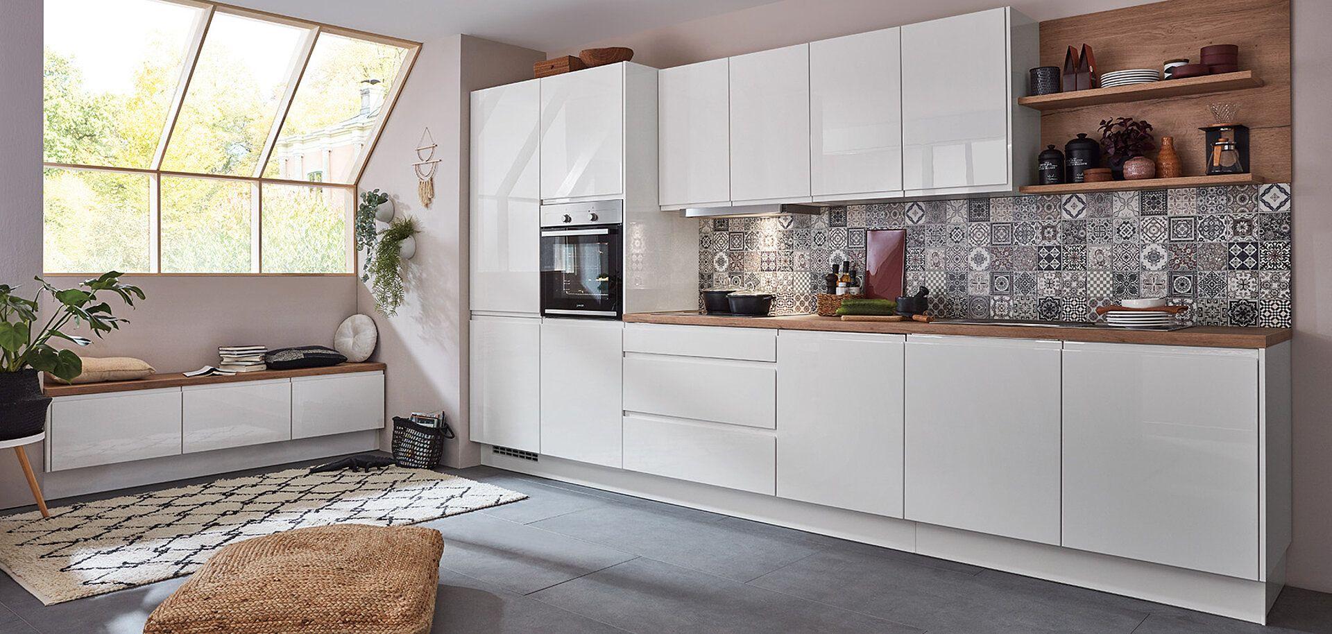 Moderne Küchenzeile mit weißen Hochglanzfronten, Holzregalen und einer dekorativen Fliesenwand. Im Vordergrund ist ein bequemer Sitzbereich und ein Teppich zu sehen, während große Fenster natürliches Licht in den Raum lassen.