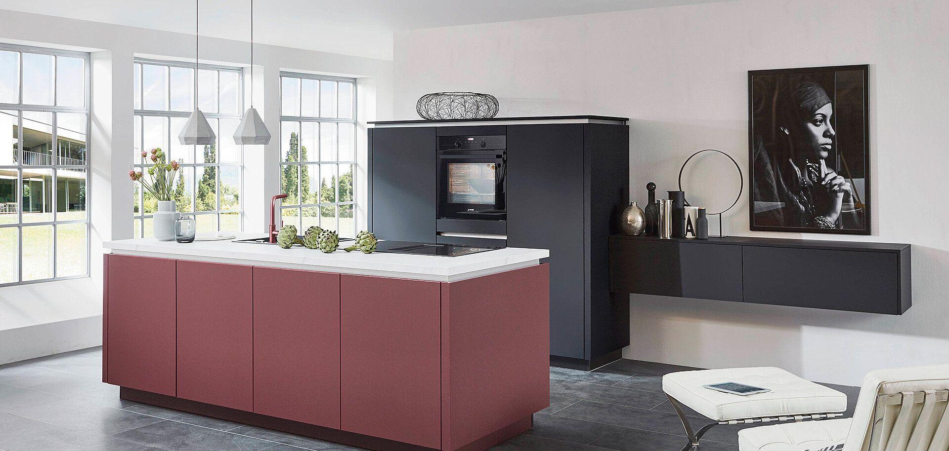 Moderne Kücheninsel in Terrakotta-Rot mit weißer Arbeitsplatte, schwarzer Küchenzeile mit integriertem Backofen und großen Fenstern in hellem Loft-Ambiente.
