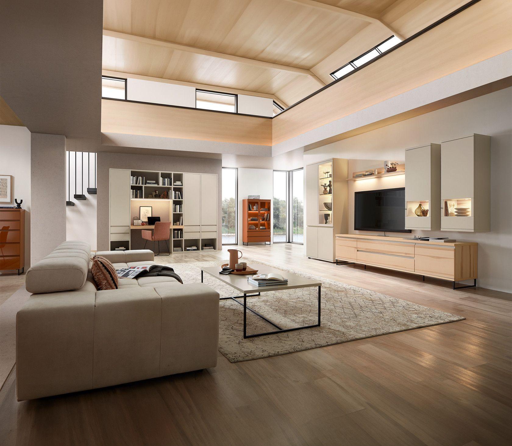 Modern eingerichtetes Wohnzimmer mit heller Wohnwand, Sofa, Couchtisch und integriertem Arbeitsplatz – großzügiger, lichtdurchfluteter Raum mit Holzdecke.
