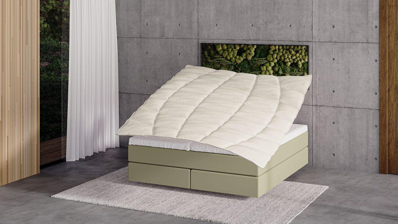 Fliegende Bettdecke über Boxspringbett in olivgrüner Farbe in modernem Schlafzimmer mit Betonwand, Moosbild als Kopfteilakzent und großem Fenster.