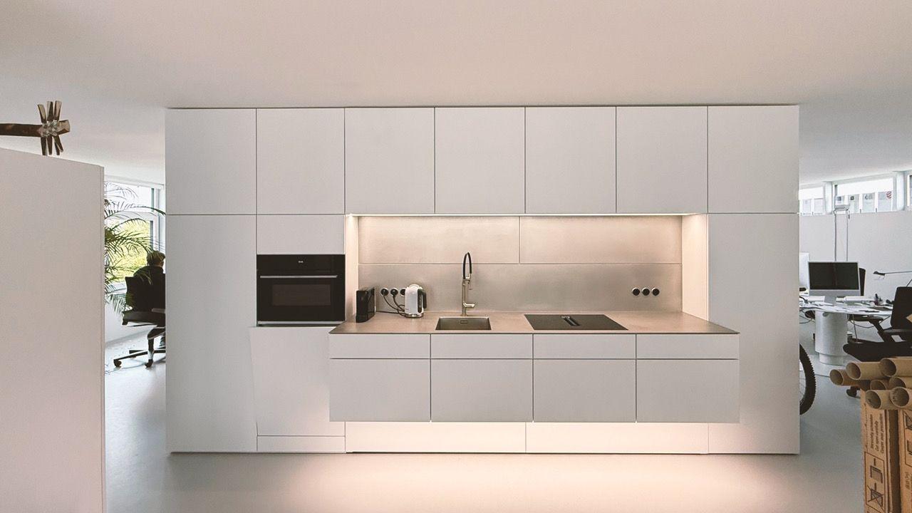 Moderne minimalistische Küchenzeile in hellem Grau mit grifflosen Fronten, integriertem Backofen, Spüle und LED-beleuchteter Rückwand in einem offenen Büro- oder Wohnraum.