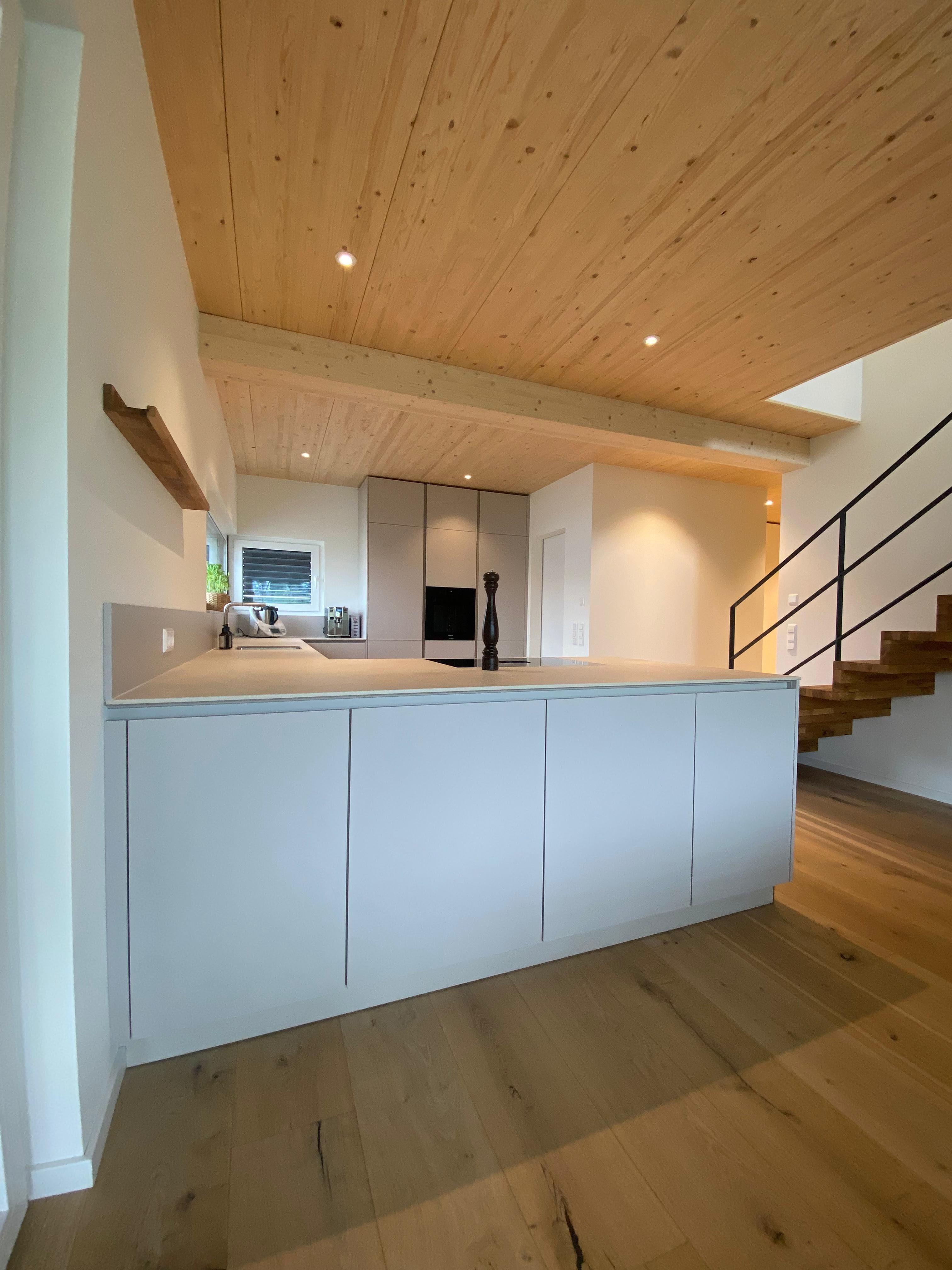 Sandfarbene Kücheninsel mit sichtbaren Holzbalken und moderner Holztreppe im Hintergrund