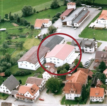 Luftaufnahme eines Gewerbegebiets mit rot umkreistem großen Industriegebäude mit rotem Dach, umgeben von kleineren Betrieben und Wohnhäusern.