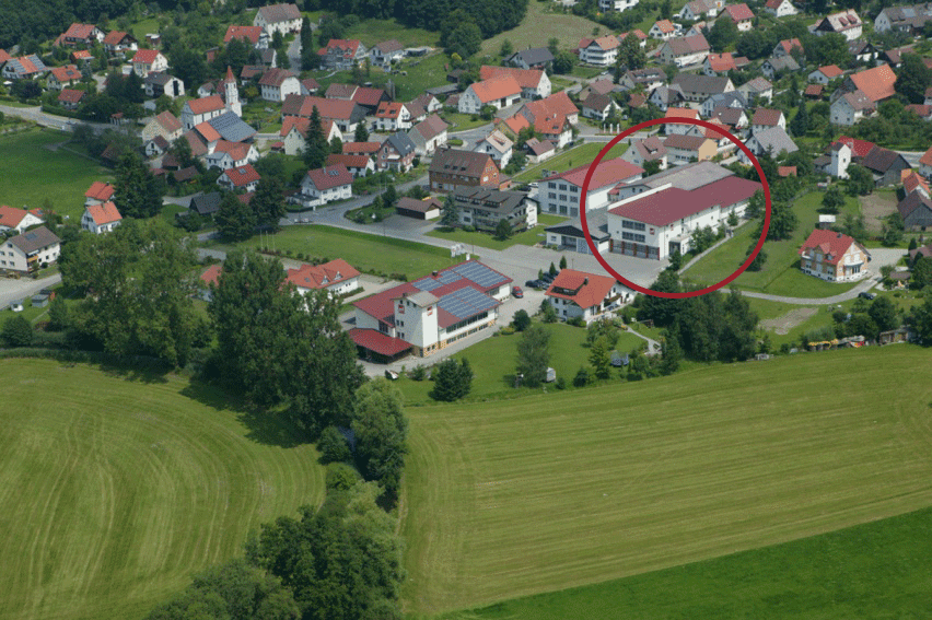 Luftaufnahme einer ländlichen Ortschaft mit rot umkreistem modernen Firmengebäude mit rotem Dach zwischen Wohnhäusern und grünen Feldern.