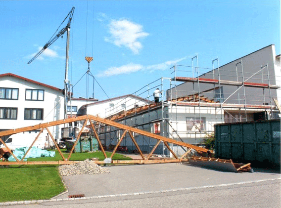 Baustelle mit Holzdachstuhl-Konstruktion und Kran vor modernen Gebäuden mit roten Dächern während der Erweiterungsarbeiten.