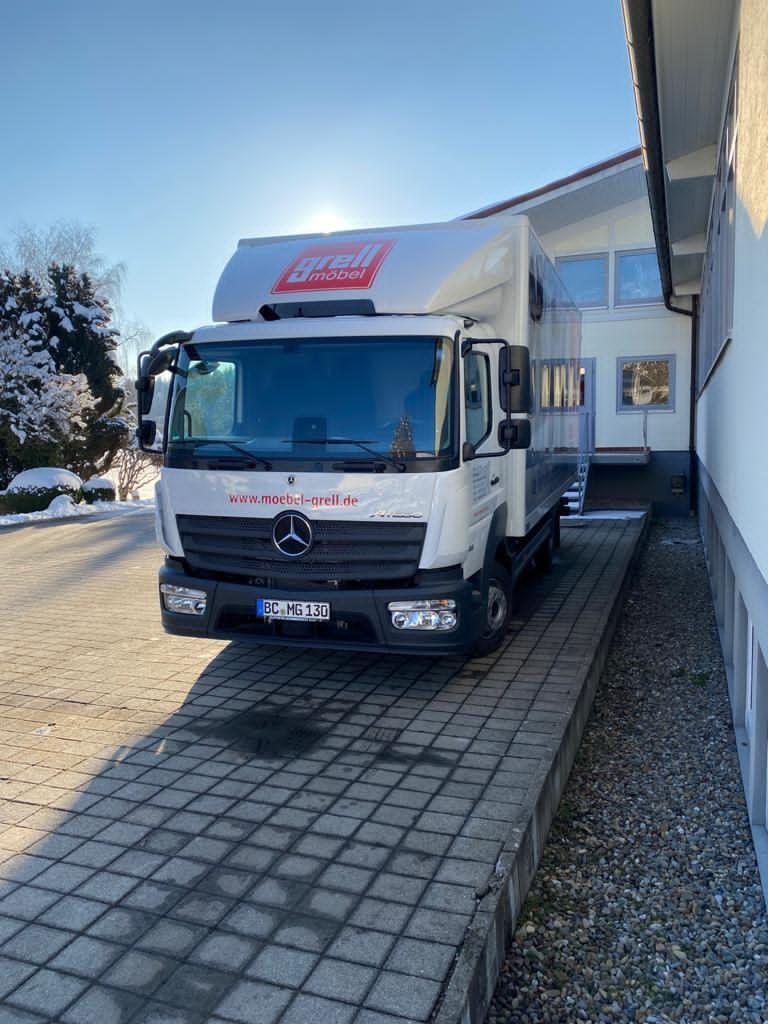 Weißer Mercedes-Lieferwagen mit "Grell Möbel" Logo steht auf gepflasterter Einfahrt vor modernem Wohnhaus im Winter.
