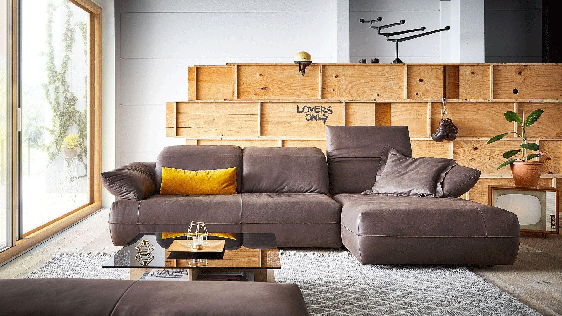 Graues Ledersofa mit Chaiselongue vor modularem Holzregalsystem, ergänzt durch gelbe Akzentkissen und modernen Couchtisch aus Holz und Glas.