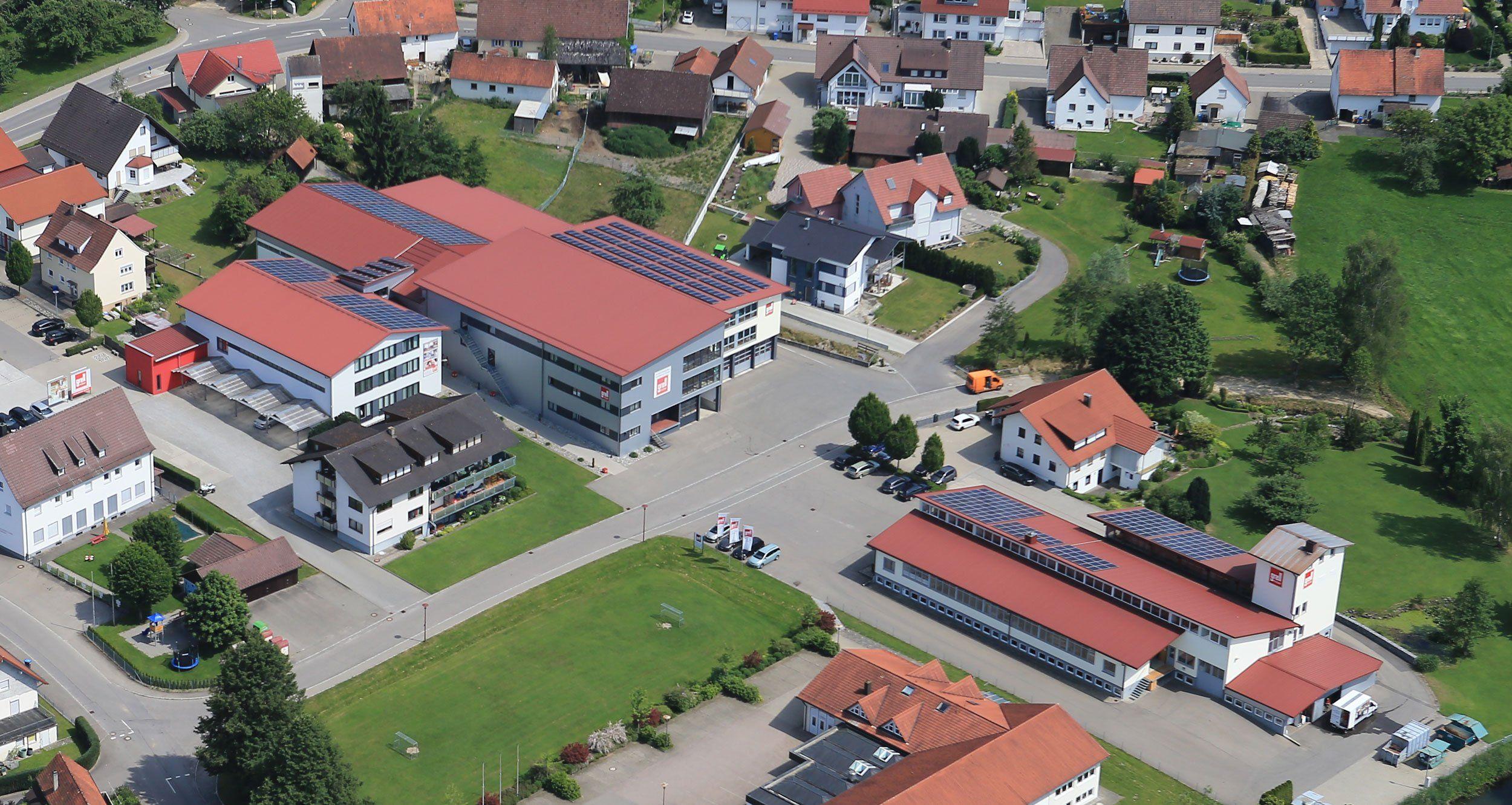 Luftaufnahme eines Gewerbegebiets mit modernen Industriegebäuden mit roten Dächern und Solaranlagen, umgeben von Wohnhäusern in ländlicher Umgebung.