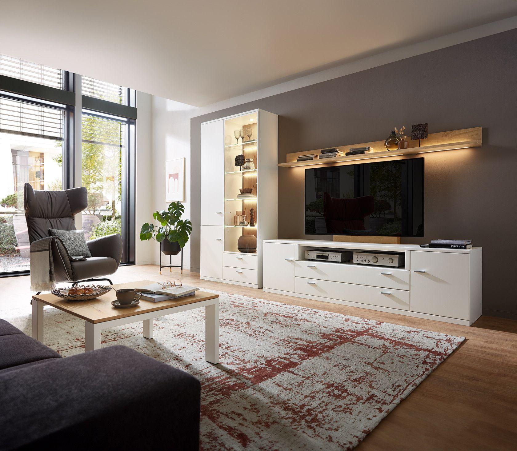 Moderne Wohnwand in Weiß mit Vitrine, TV-Element und Wandboard mit Beleuchtung – stilvoll eingerichtet in hellem Wohnzimmer mit Relaxsessel und Teppich.