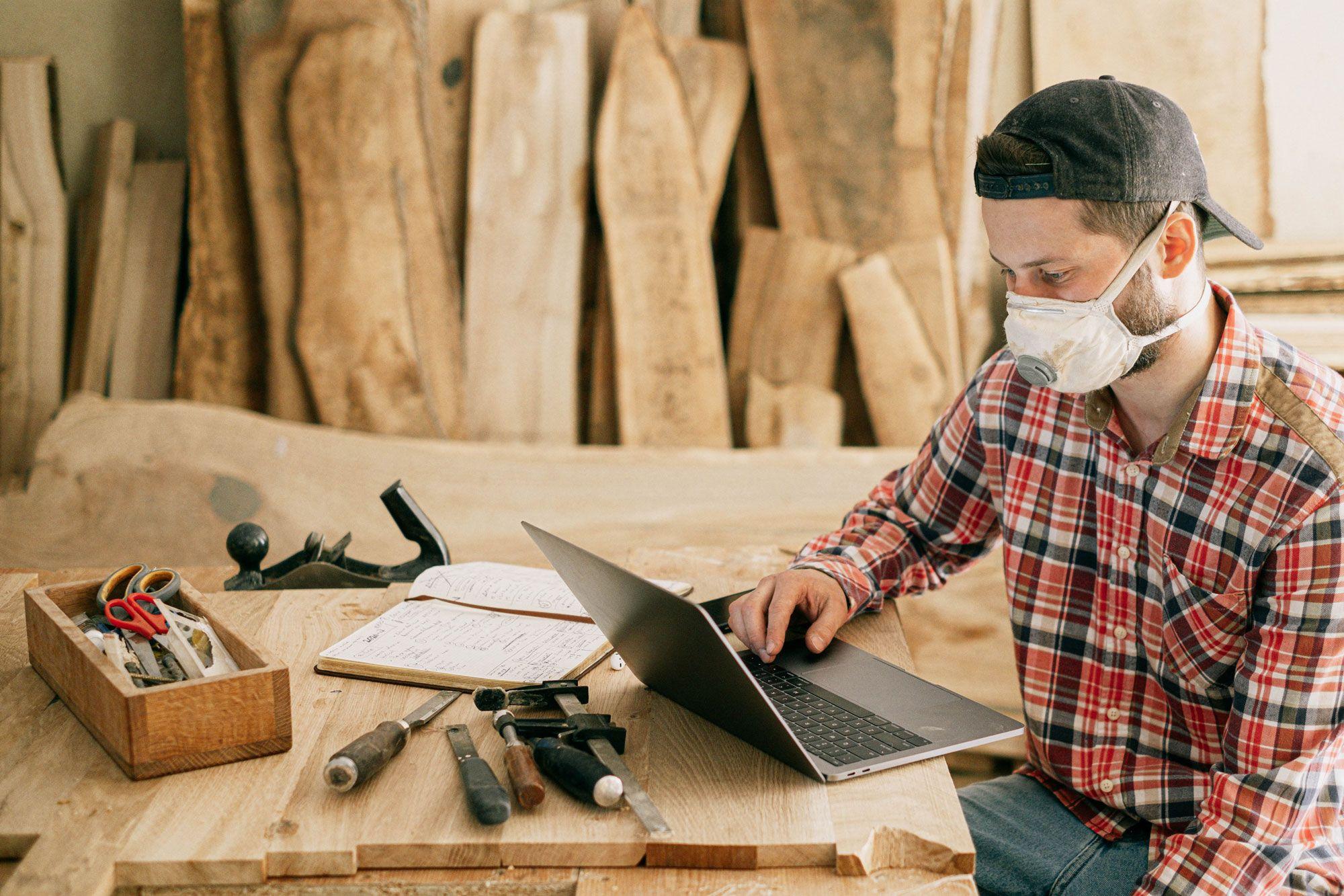 Handwerker mit Staubschutzmaske und kariertem Hemd arbeitet am Laptop in Schreinerwerkstatt, umgeben von Holzbrettern und traditionellen Werkzeugen.