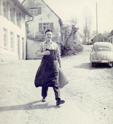 Historische Schwarz-Weiß-Fotografie zeigt einen Mann in Arbeitskleidung auf einer Dorfstraße mit alten Häusern und einem Käfer im Hintergrund.