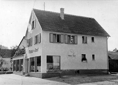 Historisches Schwarz-Weiß-Foto eines zweistöckigen Geschäftsgebäudes mit Fenstern und Eingangstür, vermutlich aus den 1950er Jahren.