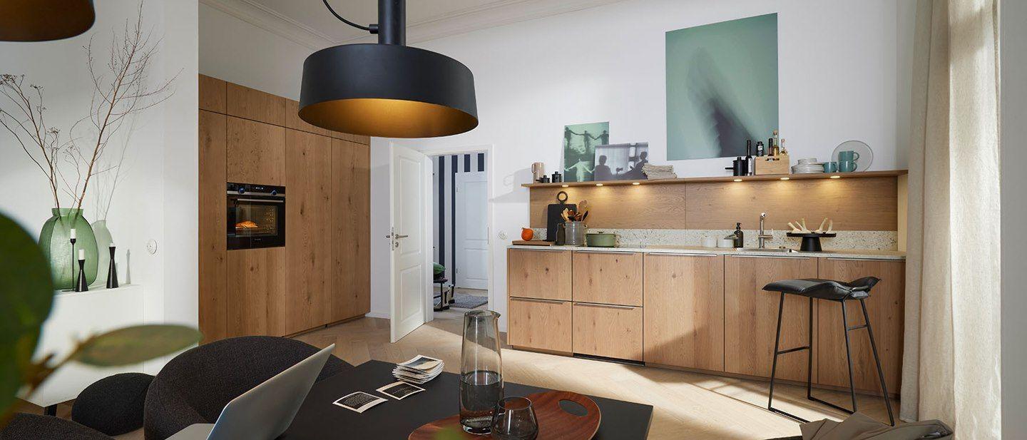 Moderne Küche in Holzoptik mit heller Arbeitsplatte, funktionalen Geräten und stilvollen Accessoires. Ein schwarzer Hängelampenschirm und eine gemütliche Sitzgelegenheit ergänzen das einladende Ambiente.