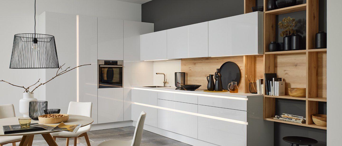 Moderne Küche in Arcticweiß mit eleganten Hochglanzfronten und einer stilvollen Holzregalwand. Die Küche ist mit einer eleganten Beleuchtung ausgestattet, die das Design betont. Ein schlichter Essbereich mit einem runden Tisch und weißen Stühlen rundet das minimalistische, funktionale Design ab.