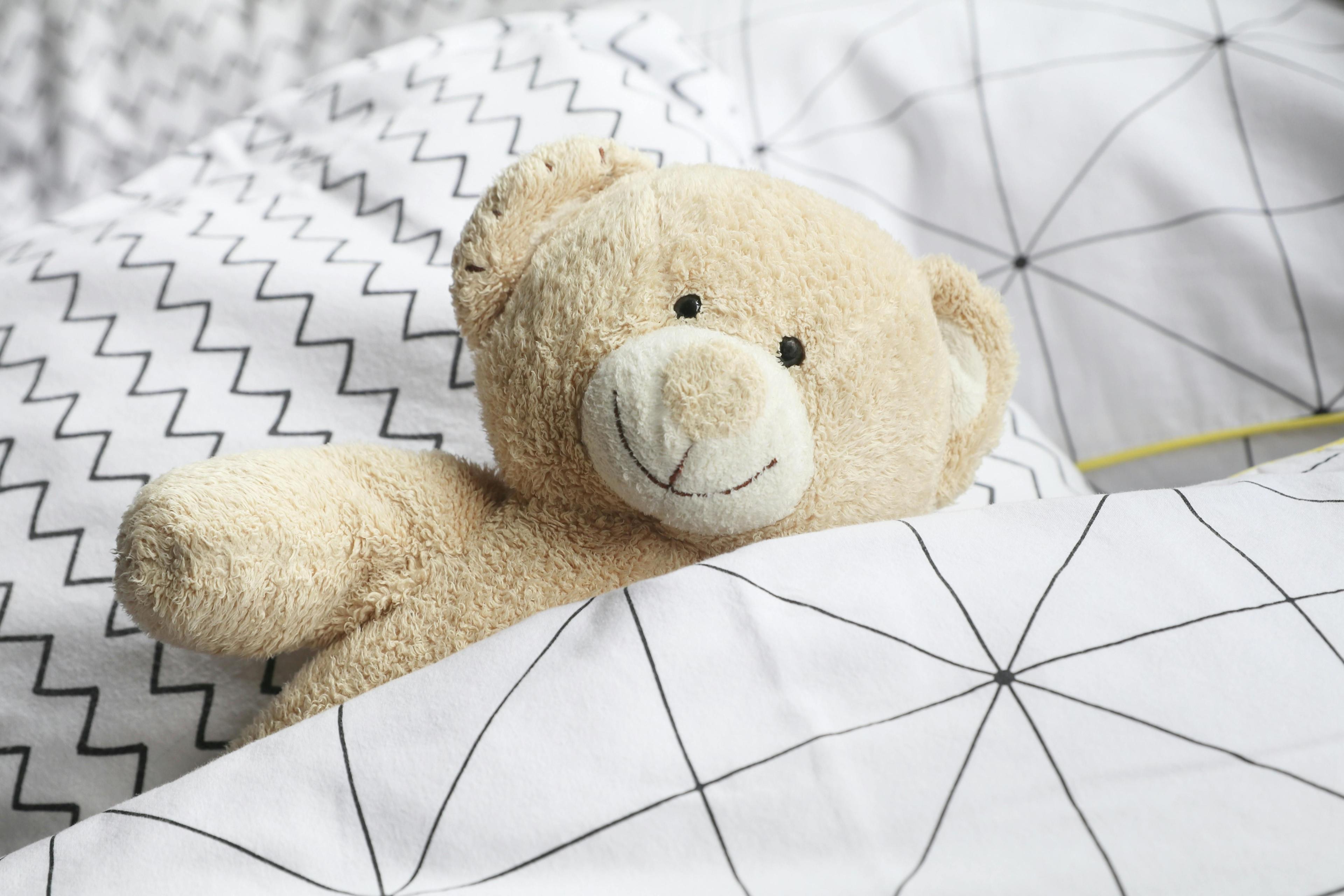 Ein beiger Teddybär liegt zugedeckt unter einer weissen Decke mit Karo-Muster