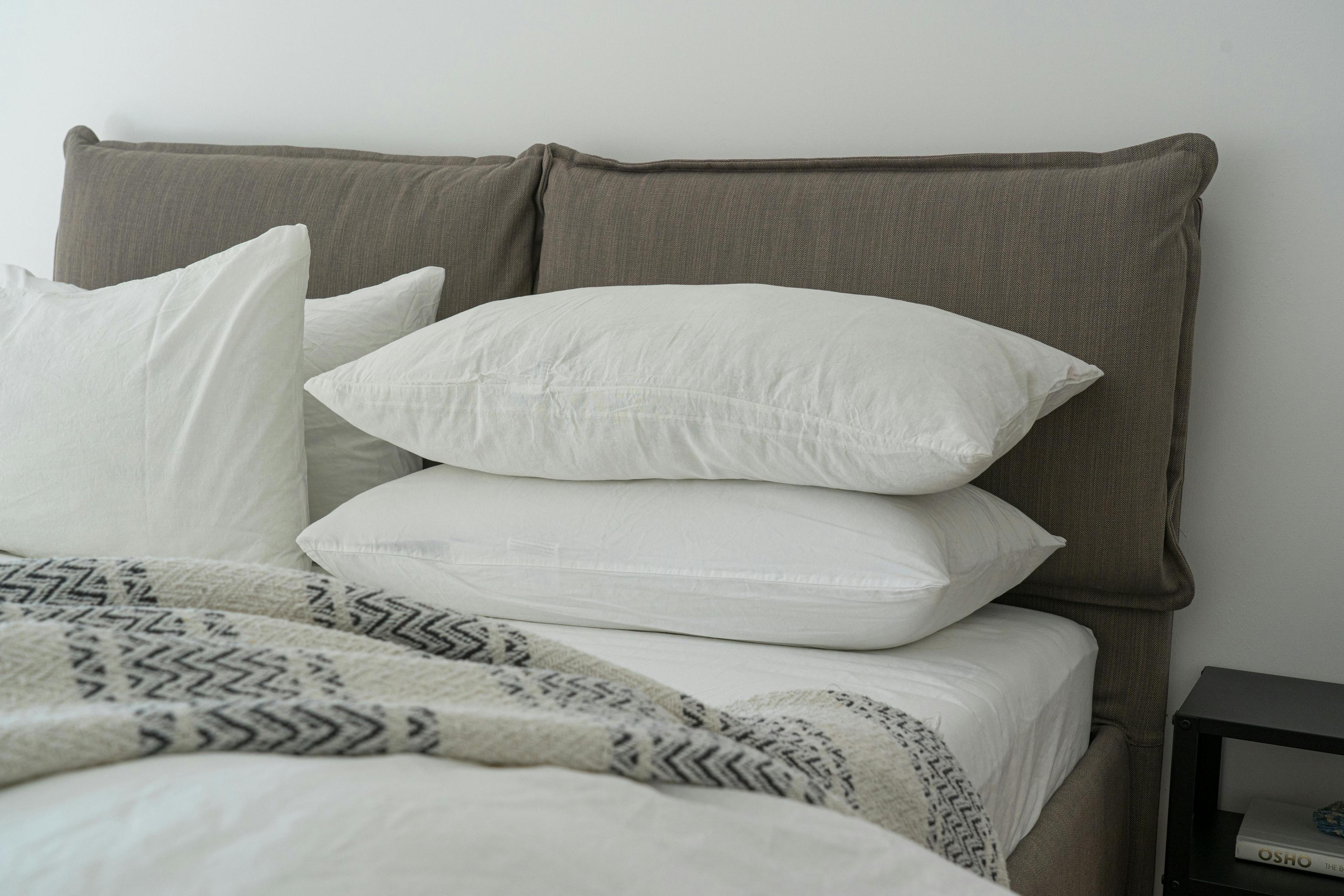 Gemütliches Bett mit grauem gepolstertem Kopfteil, weißen Kissen und einer beigen Decke mit geometrischem Muster, neben einem Nachttisch mit Büchern.