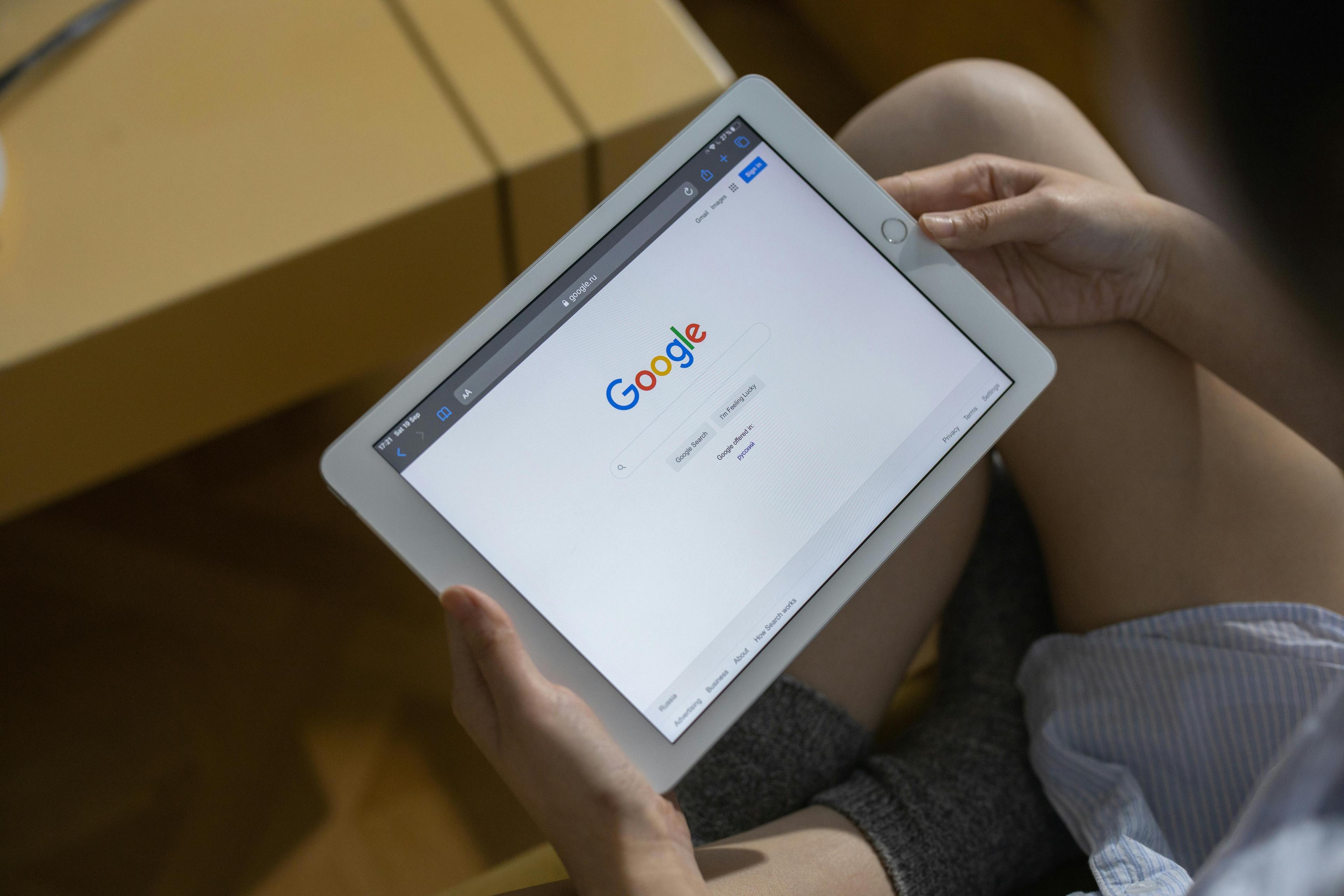 Person hält weißes Tablet mit geöffneter Google-Startseite in den Händen, sitzt entspannt auf Sofa mit Holzboden im Hintergrund.