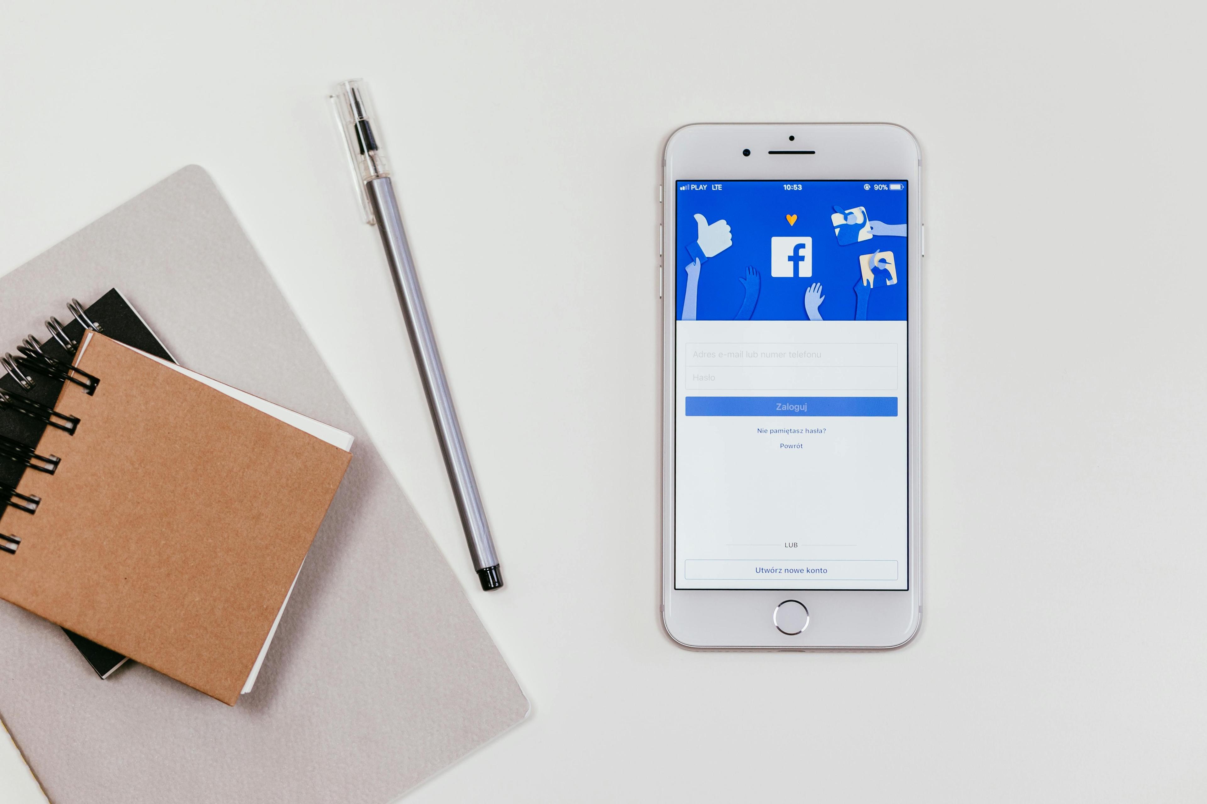iPhone mit Facebook-Anmeldebildschirm auf weißem Schreibtisch neben Notizbüchern und Stift in minimalistischer Arbeitsplatz-Anordnung.