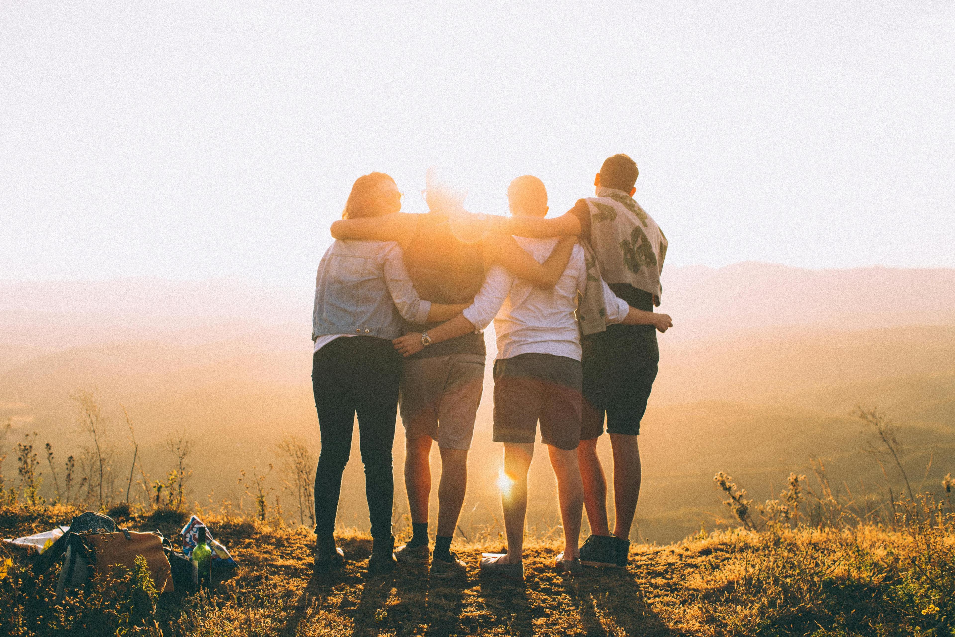Vier Personen stehen Arm in Arm im Sonnenuntergang auf einem Feld und blicken gemeinsam in die untergehende Sonne.