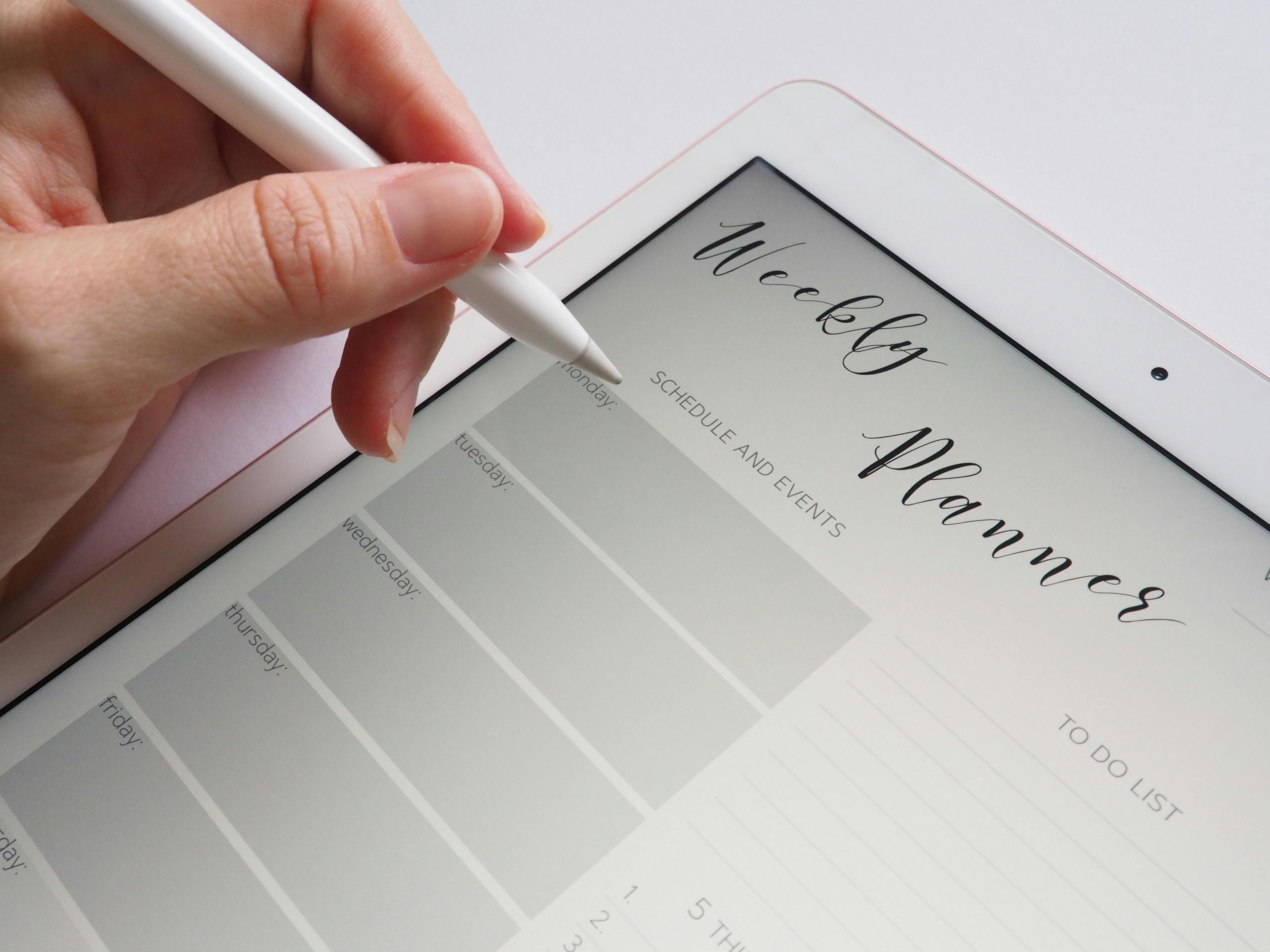 Hand mit weißem Stylus bedient digitalen Wochenplanner auf Tablet-Bildschirm, zeigt Kalenderlayout mit Wochentagen und To-Do-Liste.