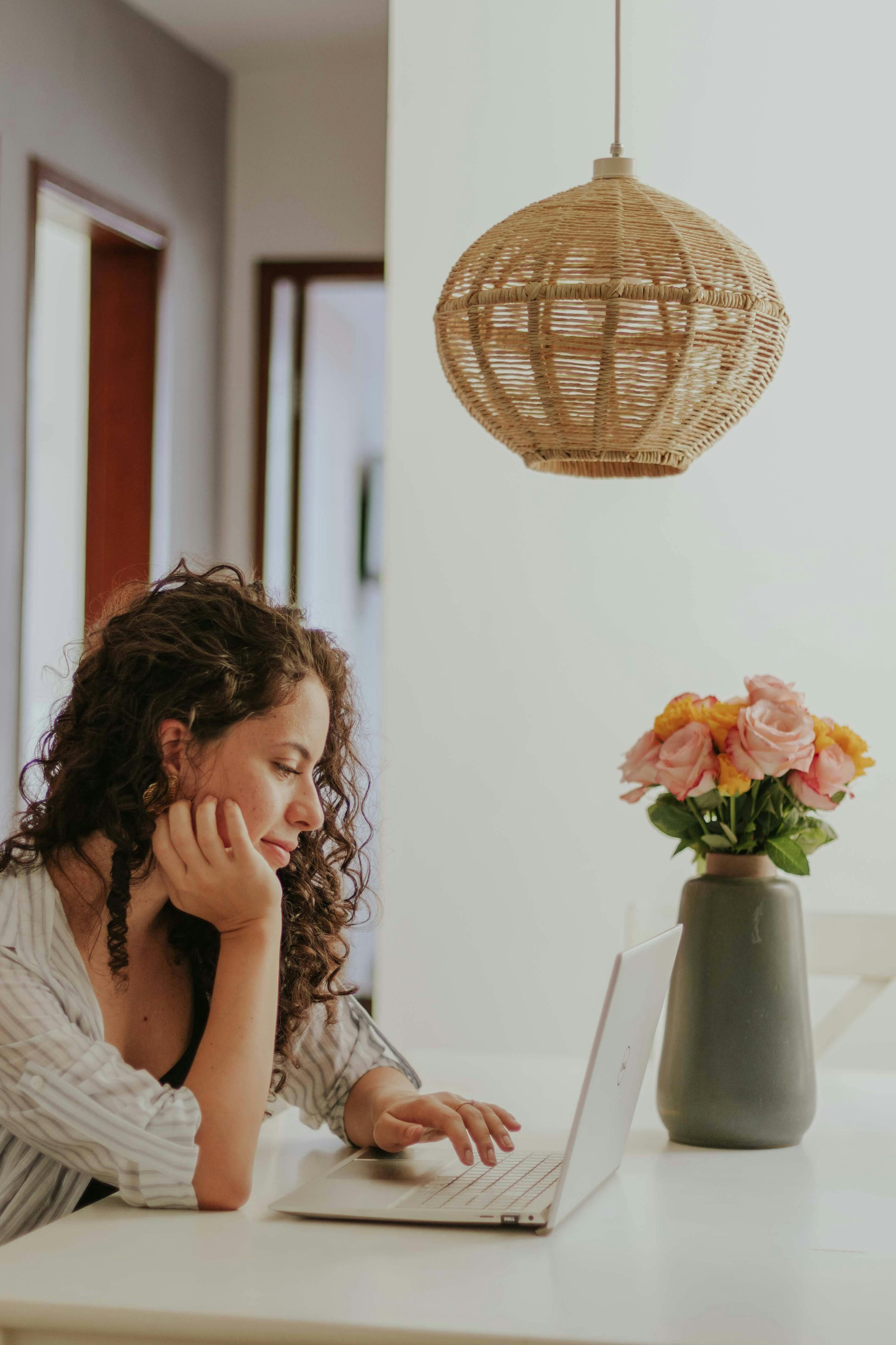 Frau mit lockigen braunen Haaren arbeitet konzentriert am Laptop an einem weißen Tisch. Rattanpendelleuchte hängt über dem Arbeitsplatz, grüne Vase mit rosa und gelben Blumen steht daneben. Moderne Wohnungseinrichtung im Hintergrund.