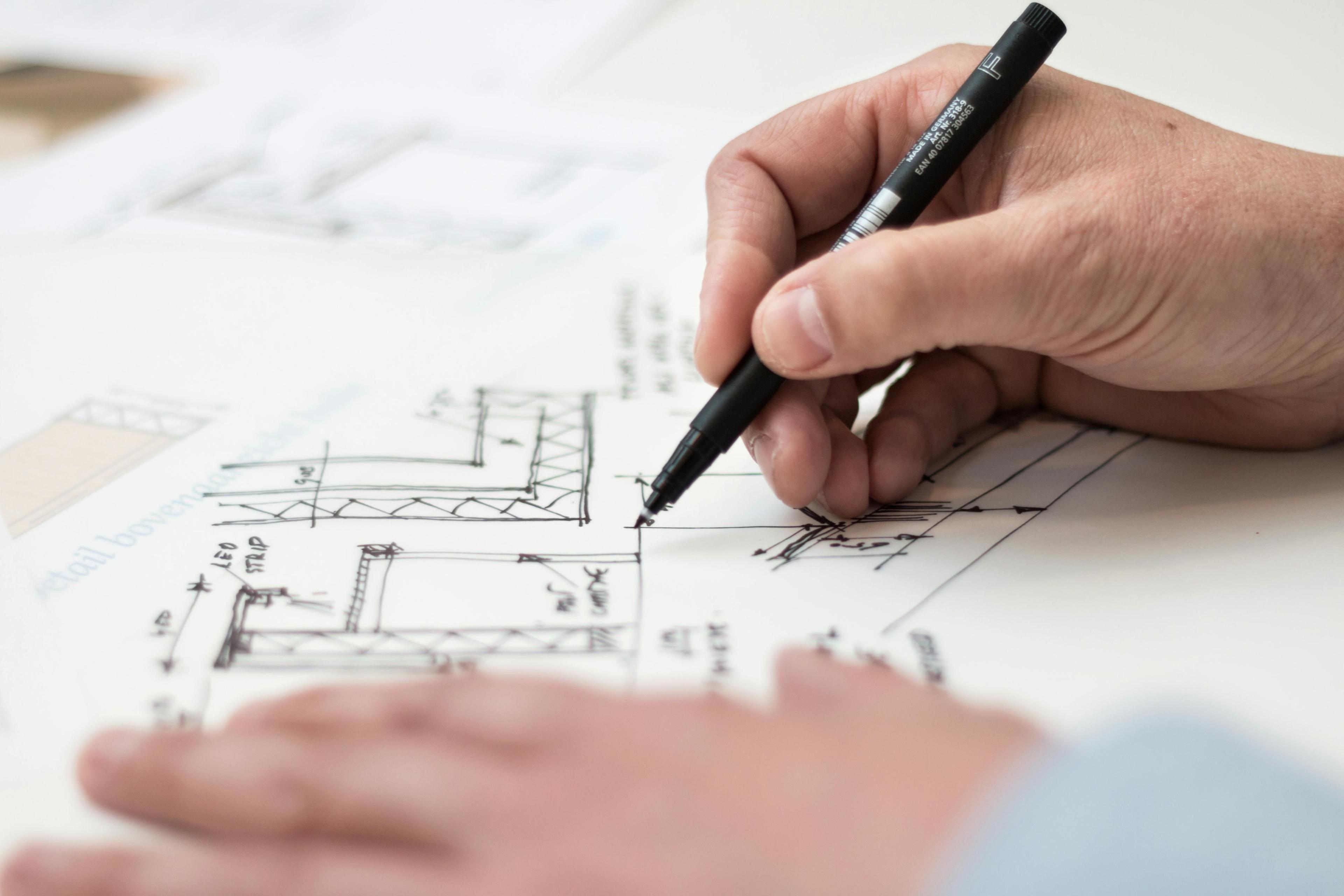 Hand mit schwarzem Stift zeichnet Details auf einen technischen Bauplan mit Maßangaben und Konstruktionszeichnungen.