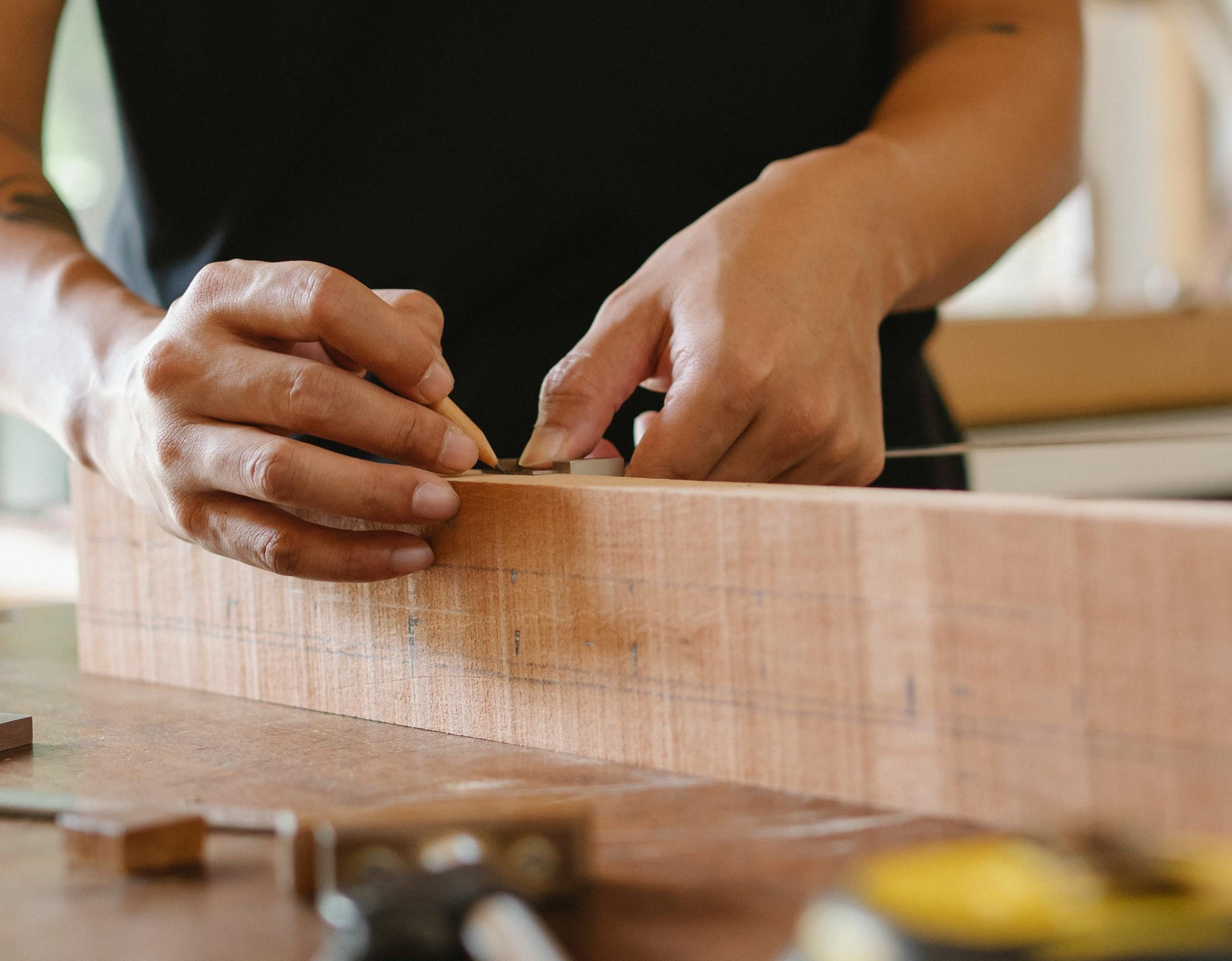 Nahaufnahme von Händen eines Handwerkers, der präzise an einem dicken Holzblock arbeitet und mit den Fingern die Oberfläche ertastet, umgeben von Werkzeugen in einer Werkstatt.