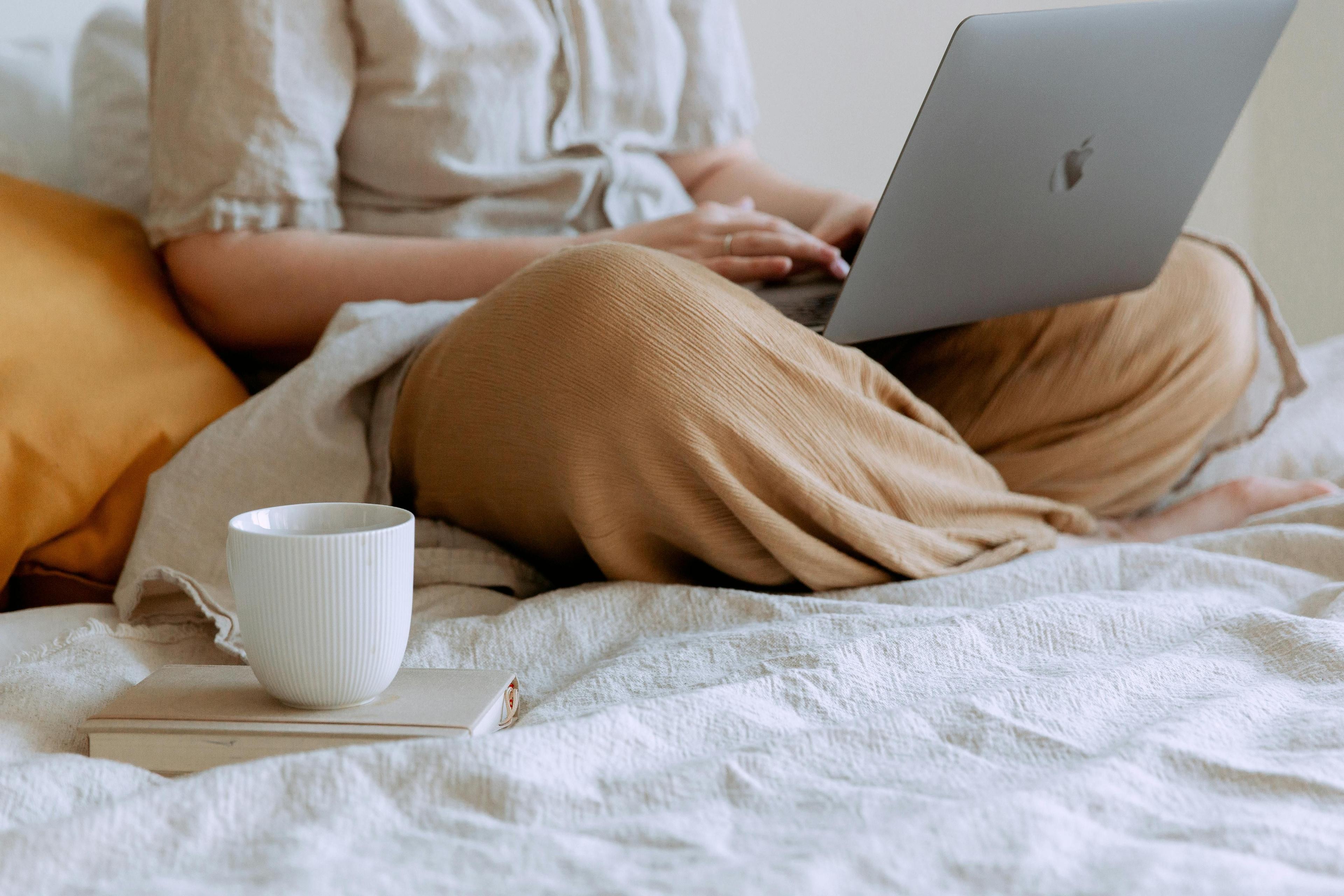 Junge Frau sitzt auf Ihrem Bett und recherchiert etwas mit Ihrem Notebook. Bei Ihr steht eine Tasse Kaffee.