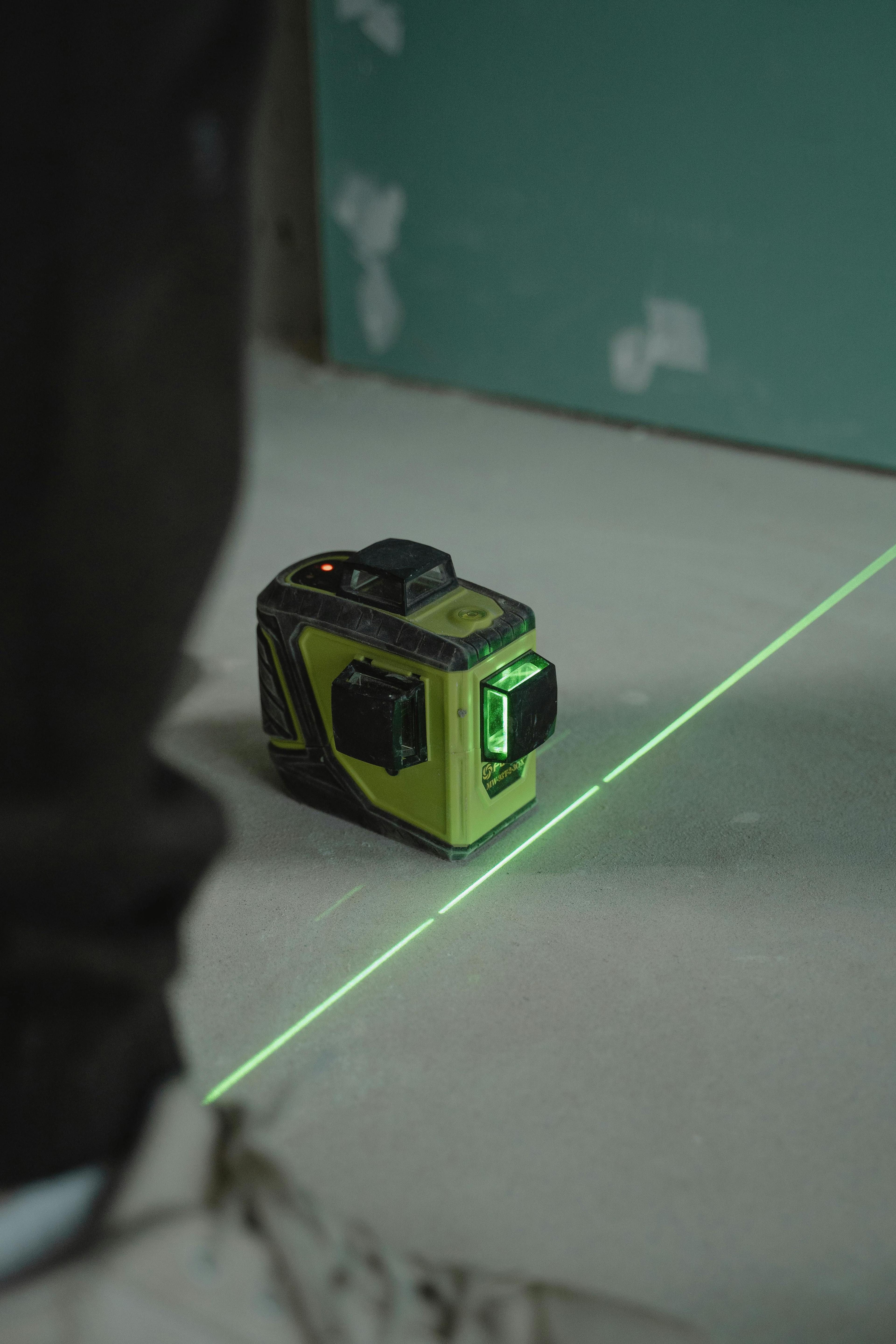 Grünes Lasermessgerät projiziert eine gerade grüne Laserlinie auf den Boden in einem Raum mit türkisfarbener Wand.