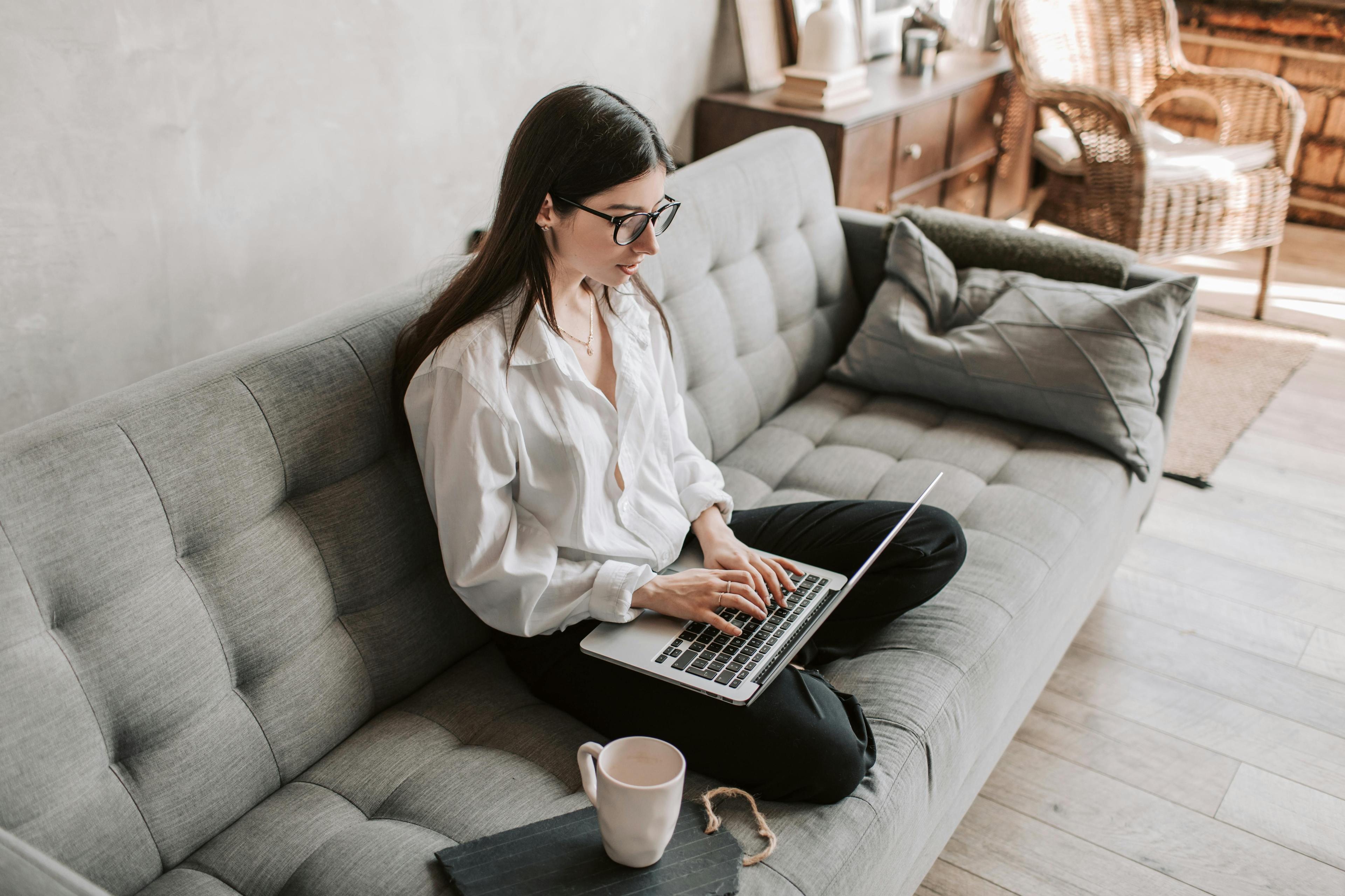 Frau mit Brille und weißer Bluse arbeitet entspannt mit Laptop auf grauem Sofa, eine Kaffeetasse steht daneben in gemütlichem Wohnzimmer.