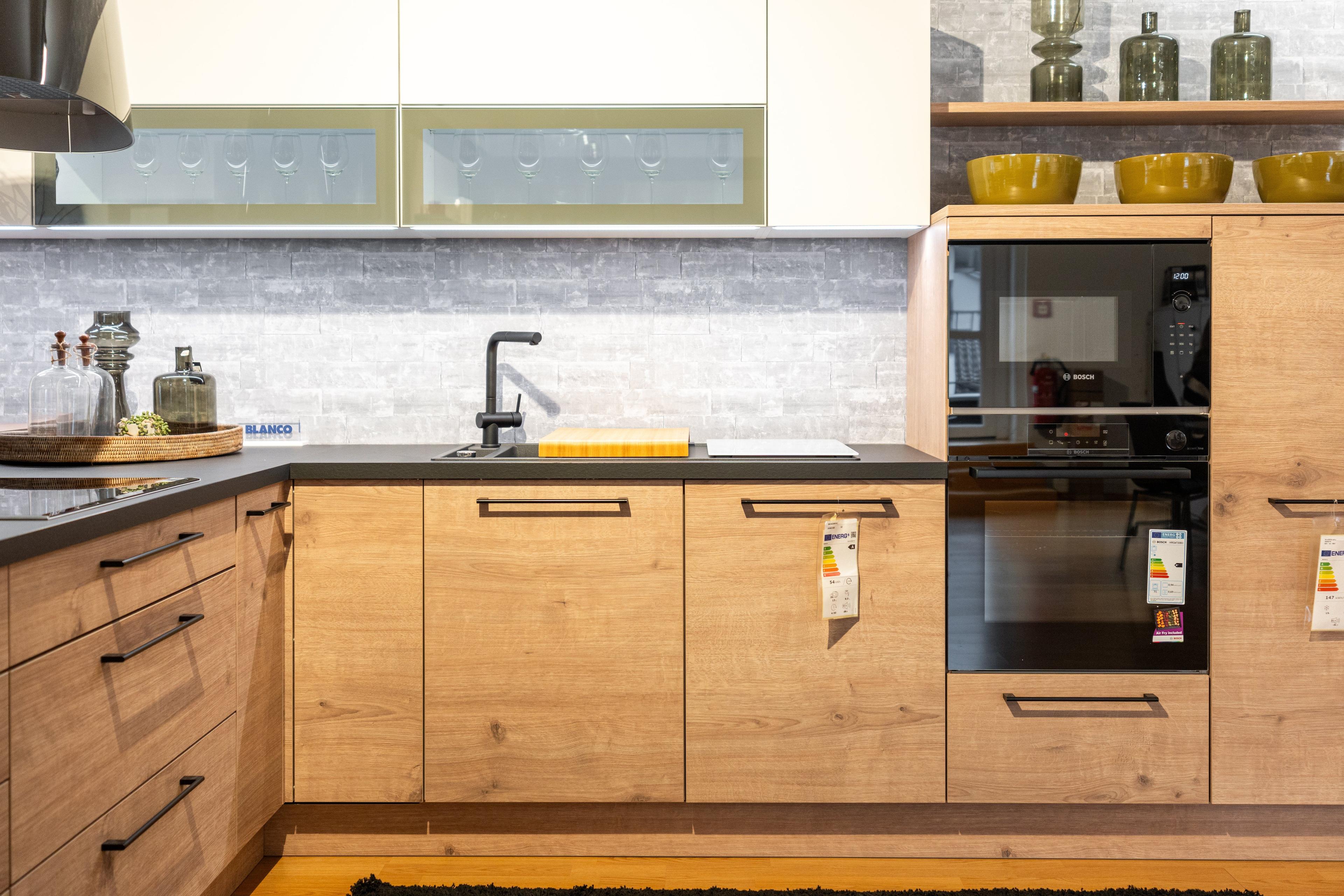 Kücheninsel mit heller Holzverkleidung und zwei grauen Barhockern mit schwarzen Metallbeinen. Schwarze Pendelleuchte darüber, graue Mosaikfliesen-Rückwand und offene Regale mit gelben Schalen.