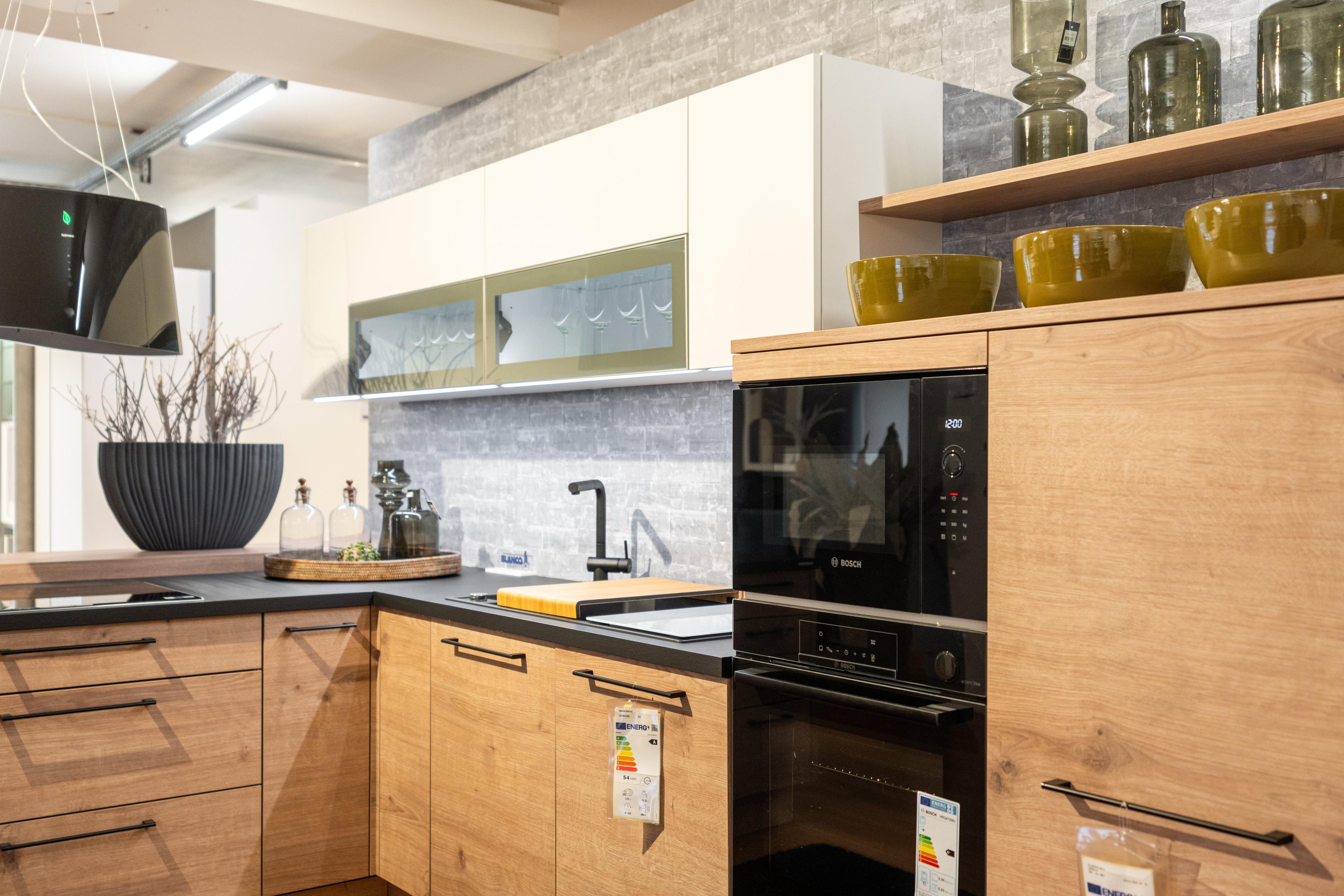 Detailansicht der Küche mit grauer Arbeitsplatte, schwarzer Spüle und Armatur, orangem Schneidebrett. Helle Holzschränke, Oberschränke mit Glasfronten und gelben Akzenten, graue Mosaikfliesen.