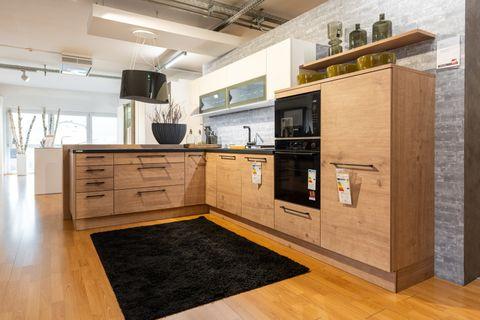 Nahaufnahme einer Küchenzeile mit hellen Holzfronten, anthrazitfarbener Arbeitsplatte und grauen Mosaikfliesen als Rückwand. Schwarze Griffe und Spüle, oranges Schneidebrett neben der Armatur.