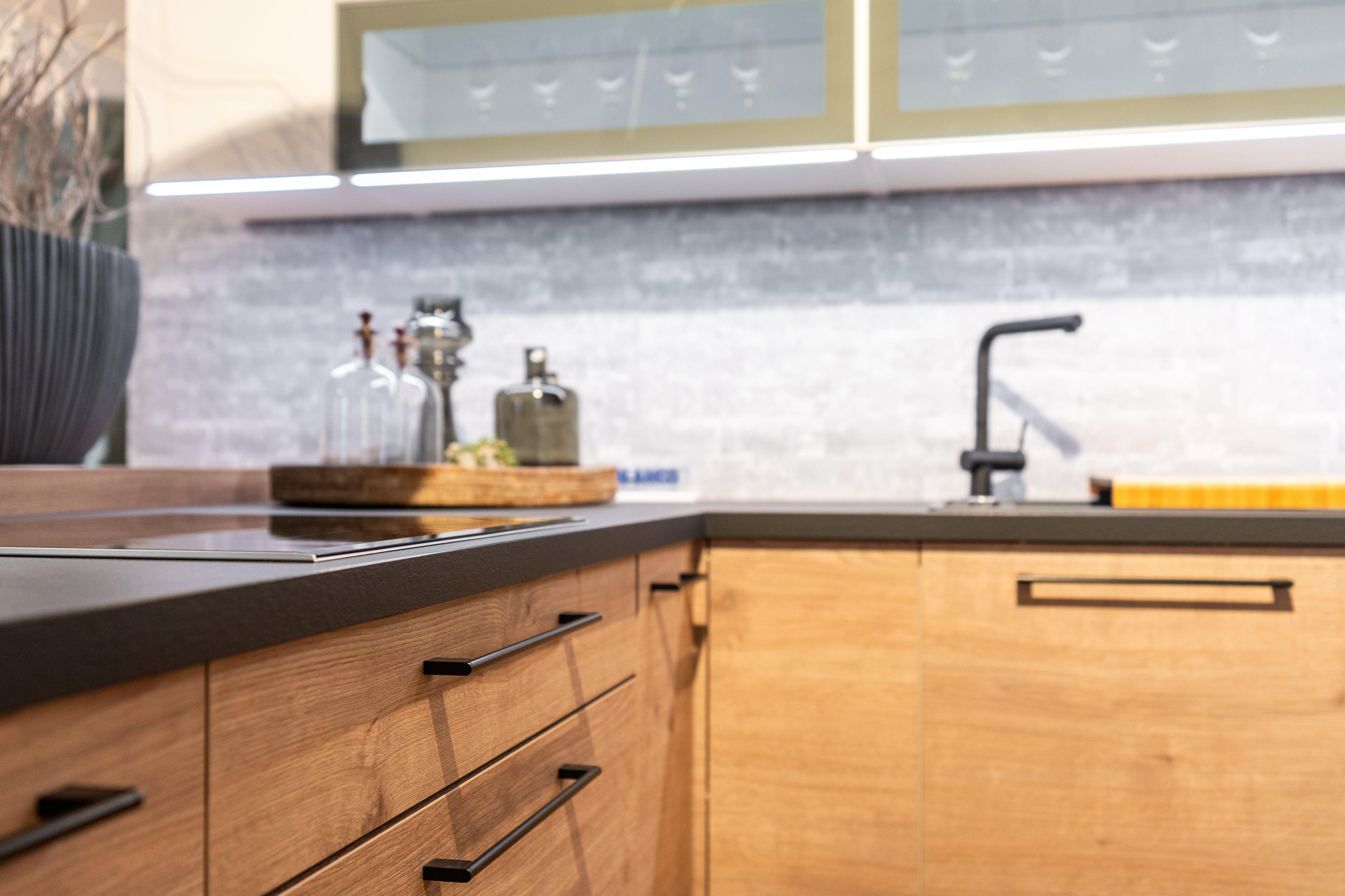 Eckbereich der Küche mit hellen Holzfronten, eingebautem schwarzem Backofen und Mikrowelle. Offene Holzregale mit gelben Schalen und grünen Glasflaschen, graue Mosaikfliesen-Rückwand.