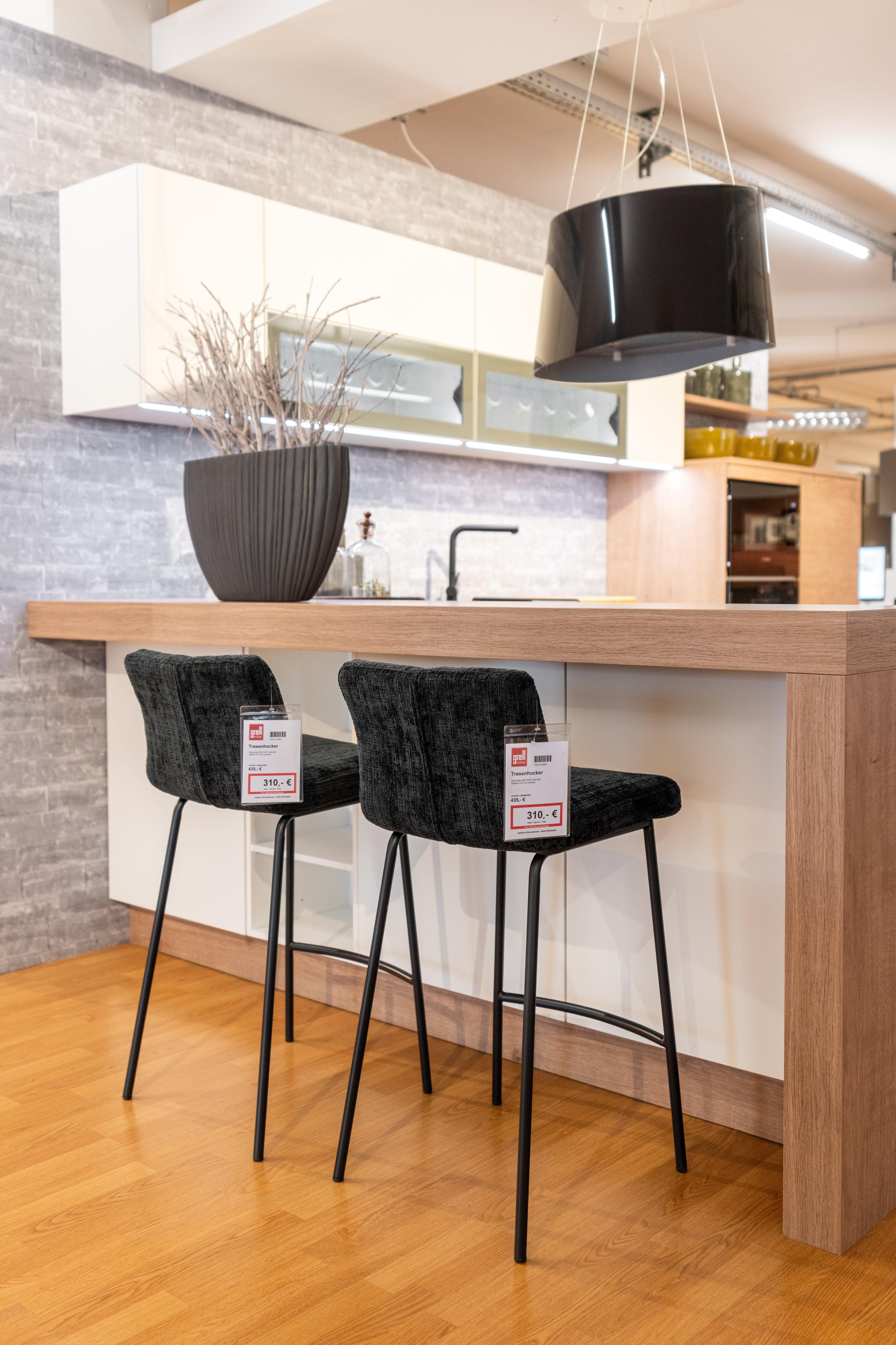 Frontale Ansicht der Küchenzeile mit hellen Holzfronten und schwarzen Griffen, graue Arbeitsplatte mit orangem Schneidebrett. Oberschränke mit Glasfronten, eingebauter Backofen rechts.