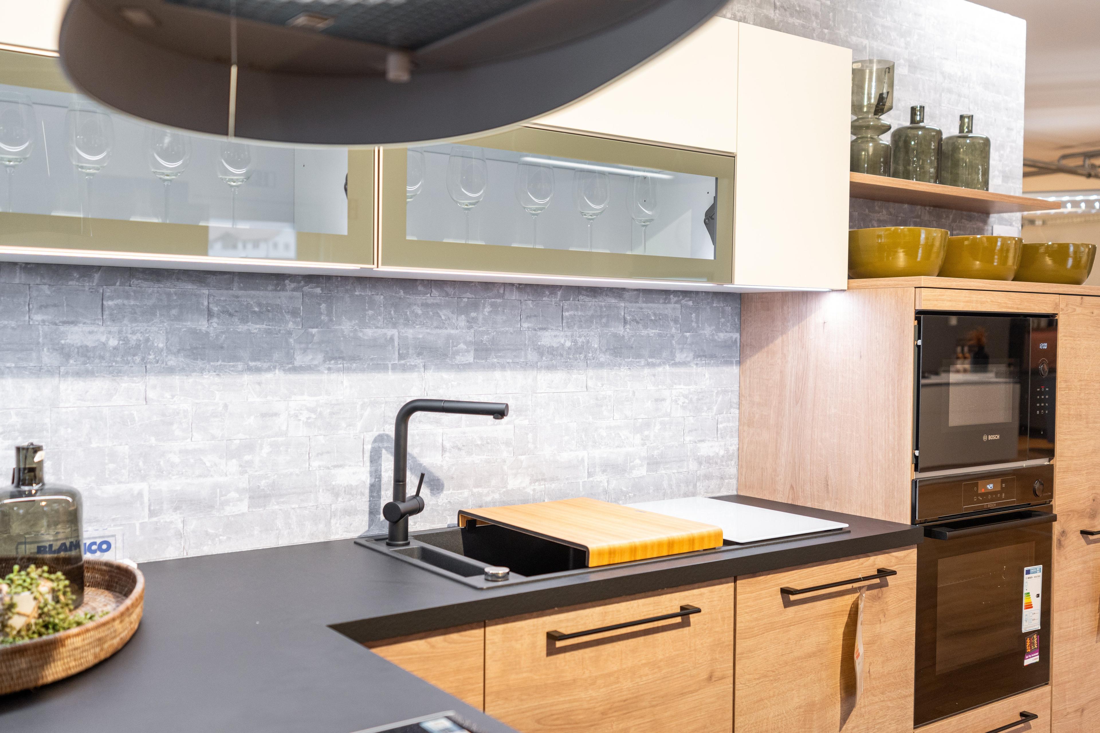 Gesamtansicht der L-förmigen Küche mit hellen Holzfronten, schwarzer Pendelleuchte und offenen Regalen. Schwarzer Teppich auf Parkett, Showroom-Ambiente mit großen Fenstern im Hintergrund.